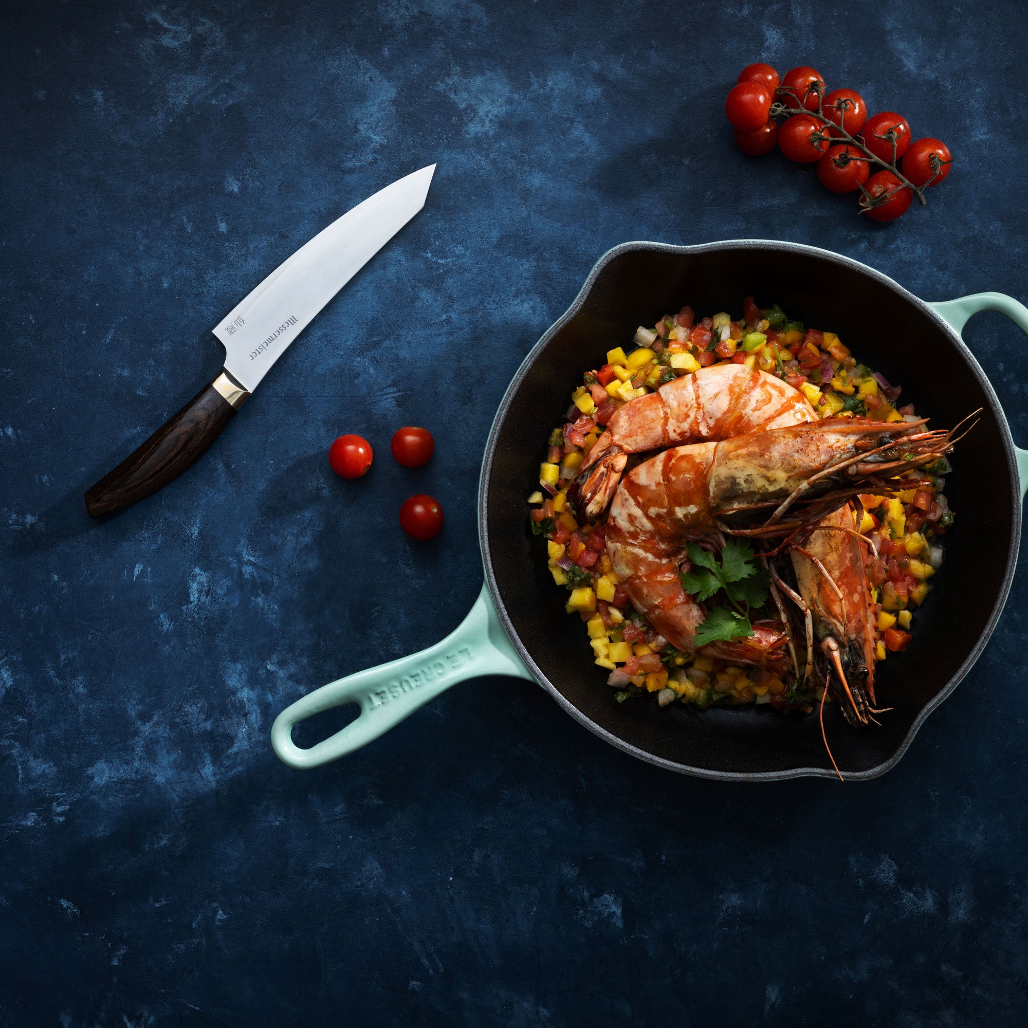 Messermeister Kawashima Utility Knife and cherry tomatoes lying next to prawns and corn sales in light blue pan on dark blue surface
