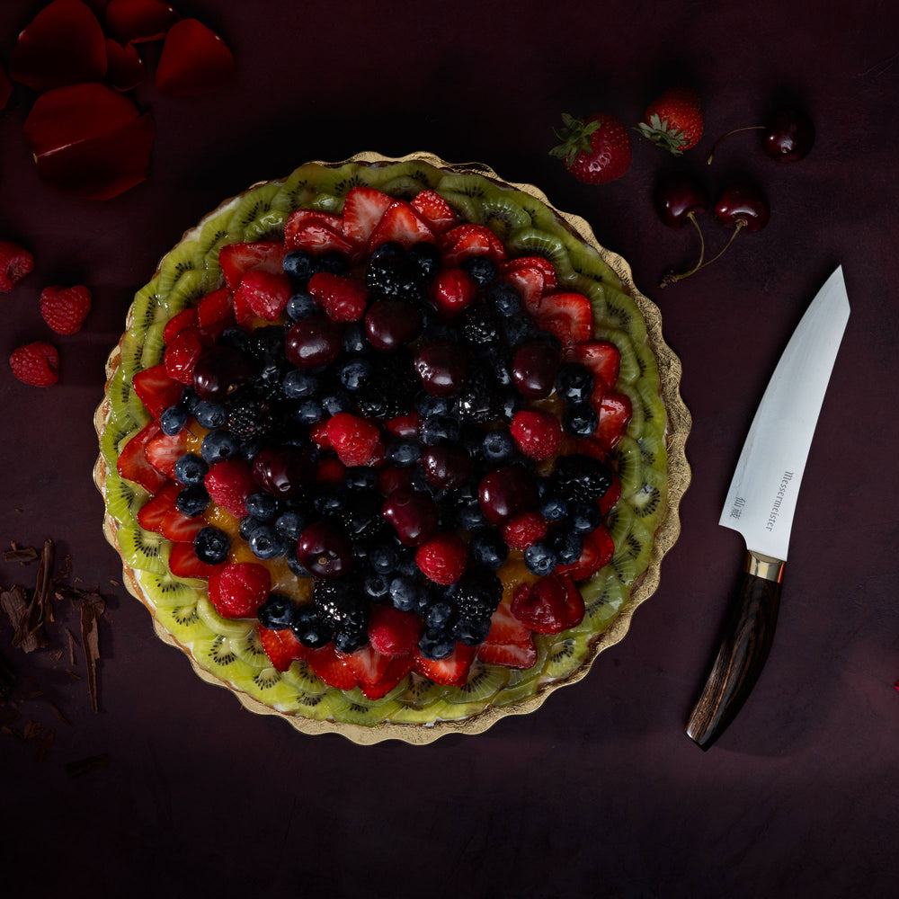 Messermeister Kawashima Utility Knife with fruit tart, cherries, shaved chocolate, strawberries and rose petals on a dark red surface