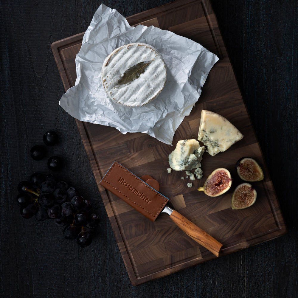 Cheeseboard with Brie, blue cheese, figs, and grapes on a wooden board with a Messermeister Oliva Paring Knife sheathed by a Messermeister Magnetic Leather Edge Guard in Saddle Color