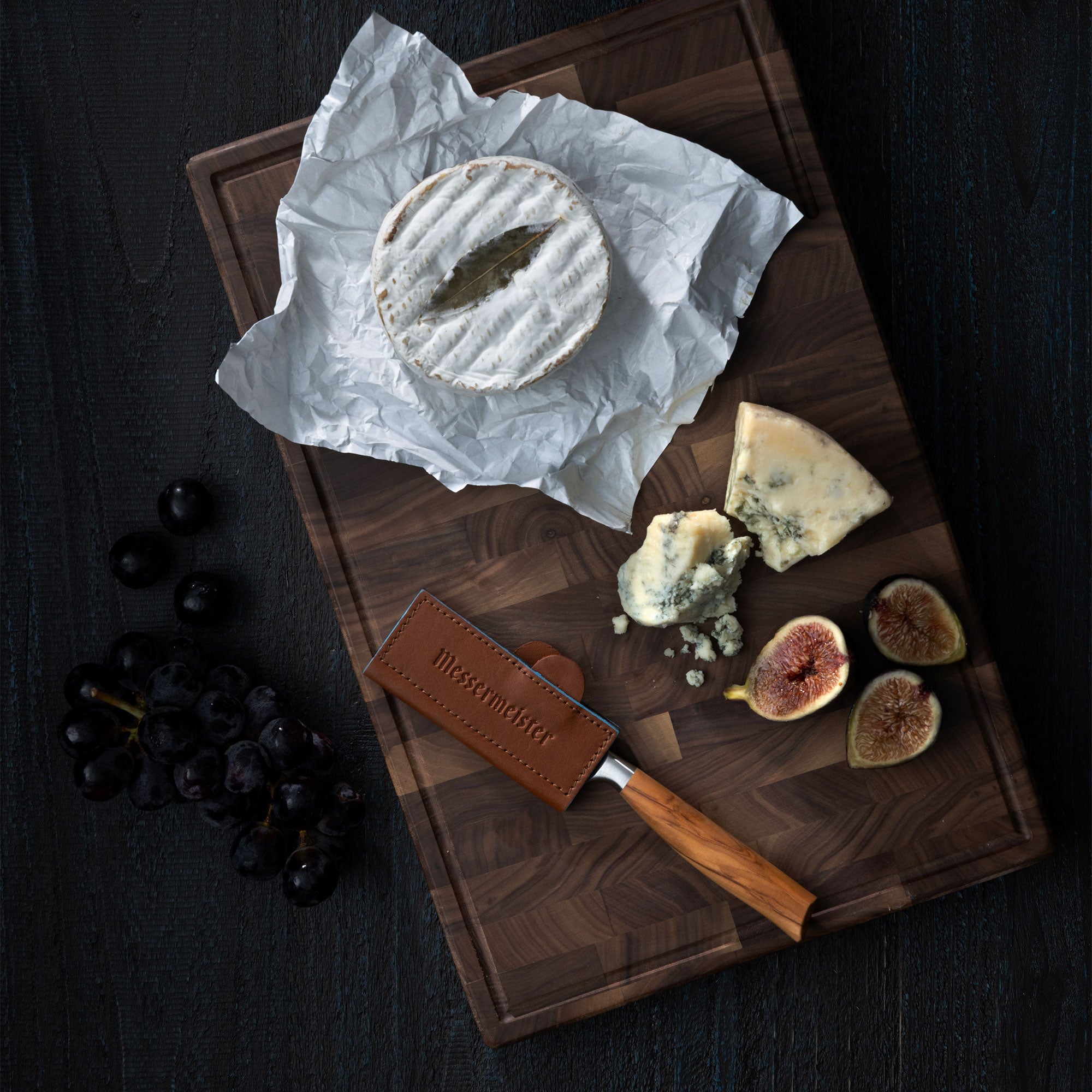 Cheeseboard with Brie, blue cheese, figs, and grapes on a wooden board with a Messermeister Oliva Paring Knife sheathed by a Messermeister Magnetic Leather Edge Guard in Saddle Color