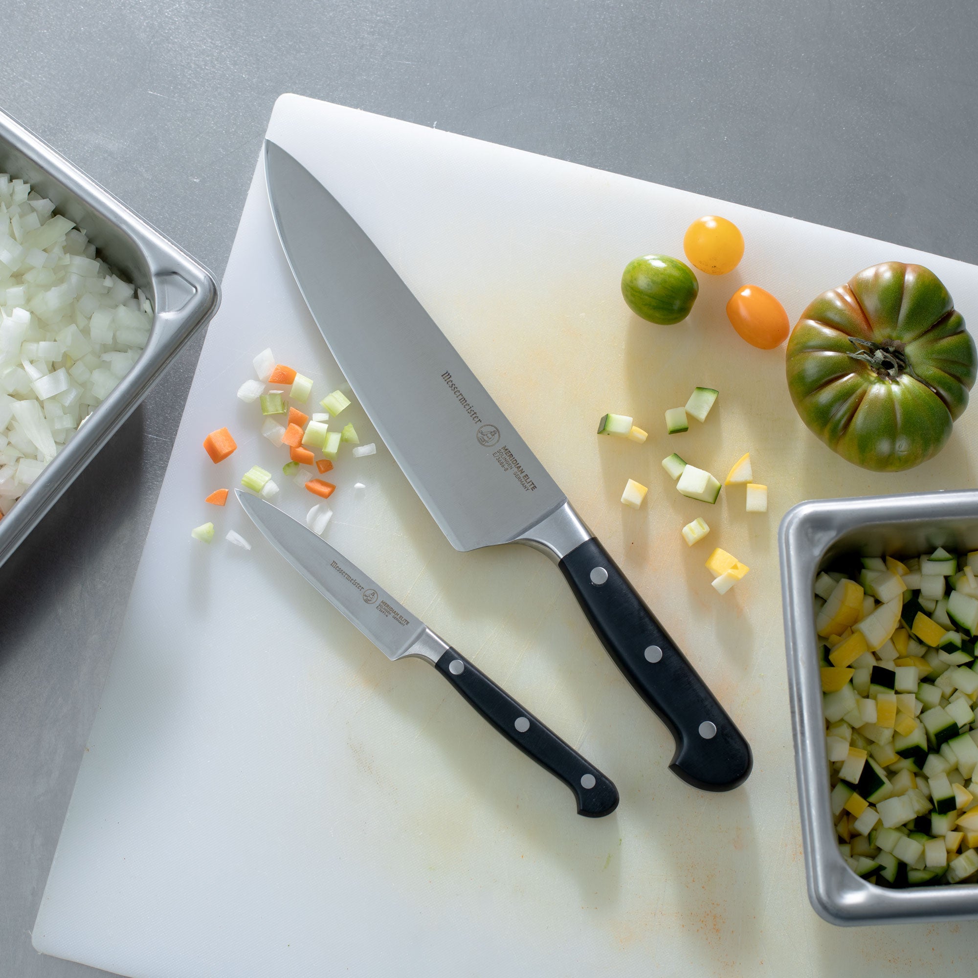 Messermeister Meridian Chef and Paring Knife on White Cutting Board on Stainless Steel Prep Station with chopped vegetables.