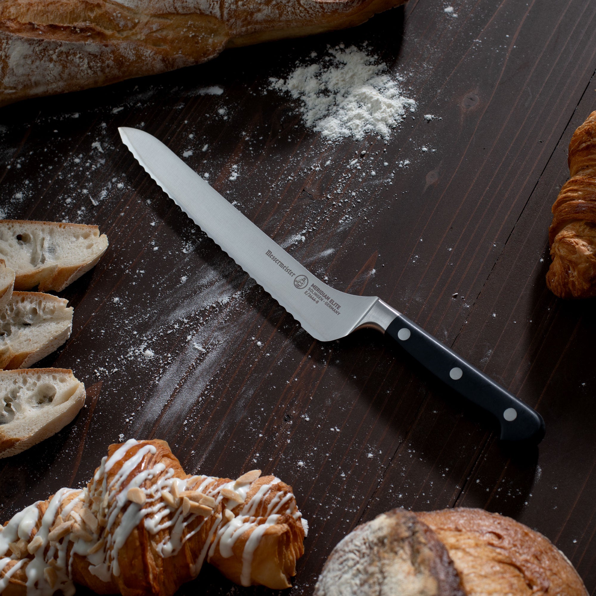 Messermeister Meridian Offset Scalloped Bread Knife on a wooden surface with bread slices and pastries