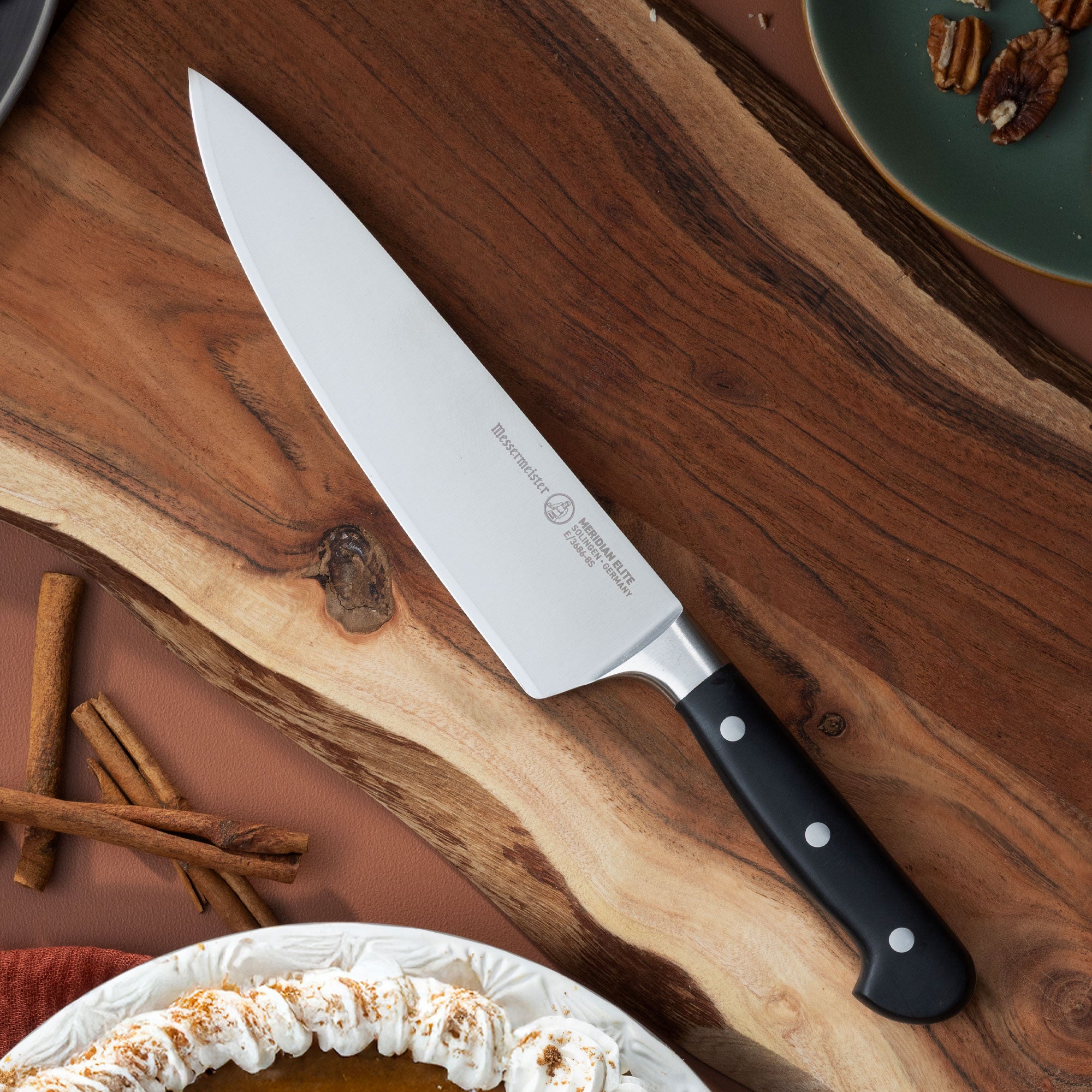 Messermeister Meridian Chef Knife with a black handle on a wooden surface with cinnamon sticks and a pie.