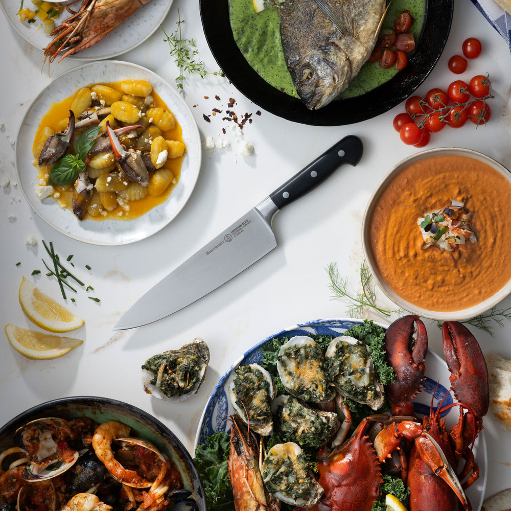 Assorted seafood dishes including lobster, clams, and oysters on a white table with a Messermeister Merdiain Chef's knife.