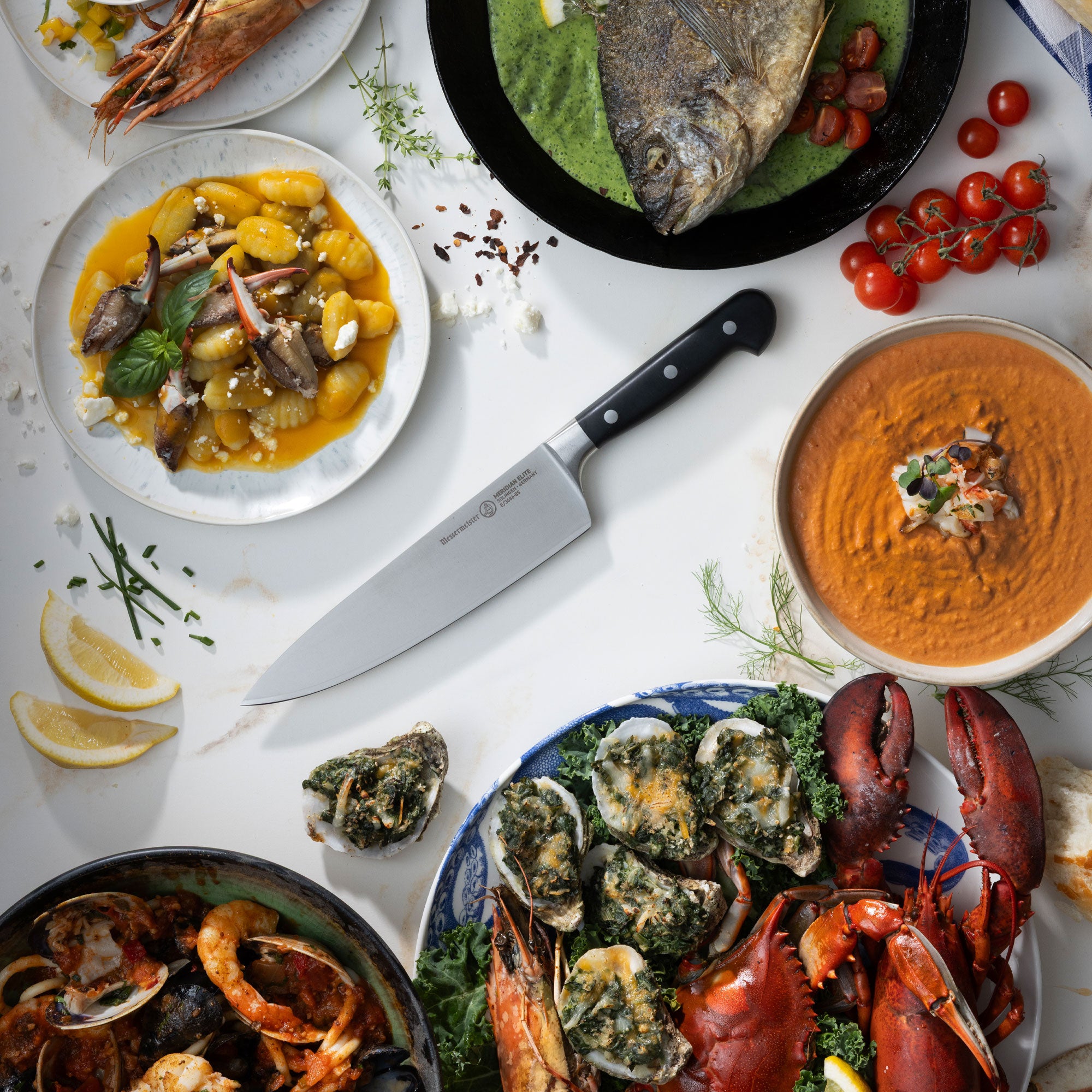 Assorted seafood dishes including lobster, clams, and oysters on a white table with a Messermeister Merdiain Chef's knife.