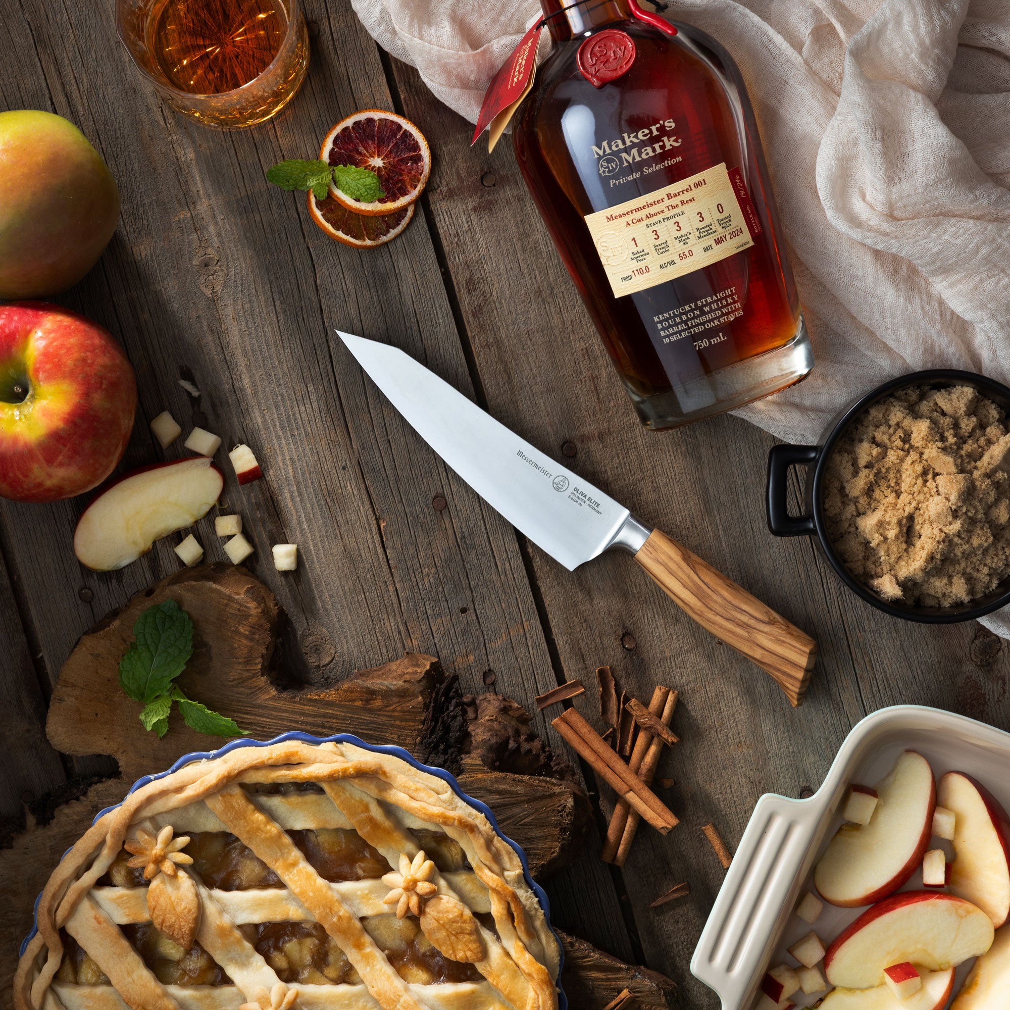 Maker's Mark bourbon bottle with pie, apples, and spices on a wooden surface with Messermeister Oliva O.T. Prep Knife.
