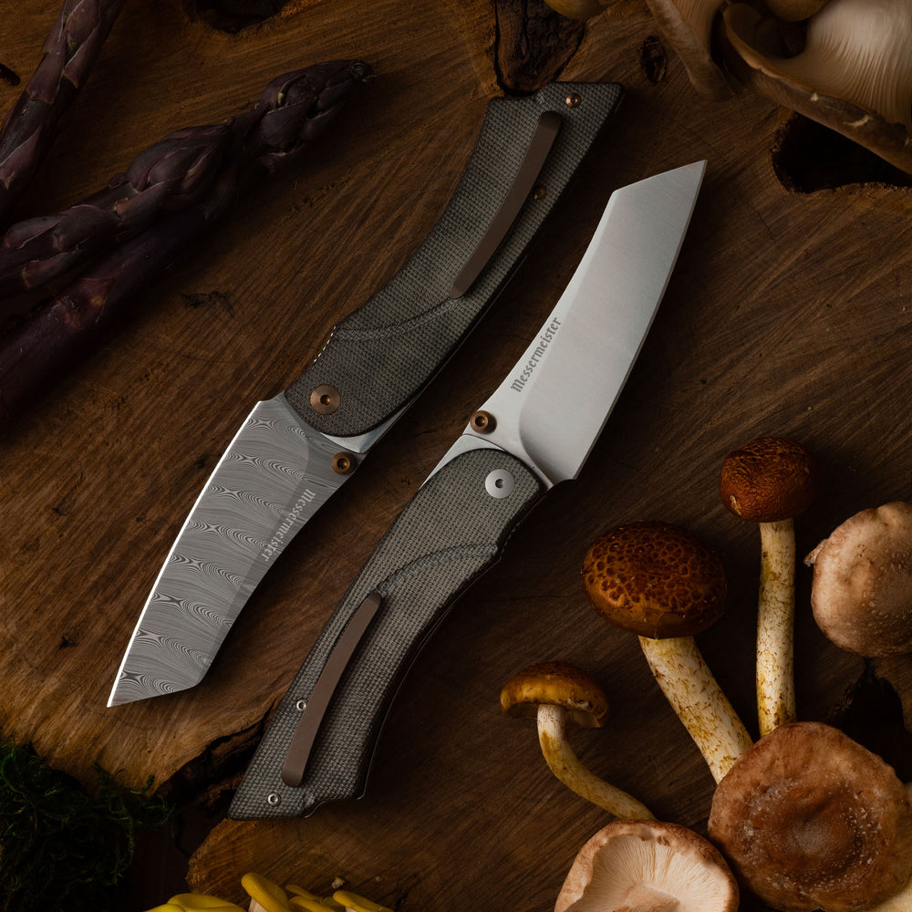 Messermeister Ojai Folding knives with in stainless steel and damascus variation on a wooden surface with mushrooms and asparagus.