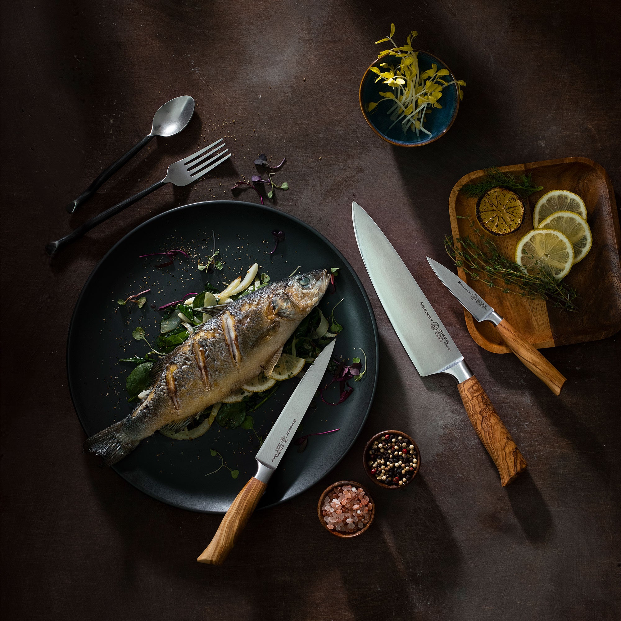 Fried fish on a dark plate with knives, lemon slices, and herbs on a wooden surface featuring the Messermeister Oliva Chef, Utility and Paring Knife