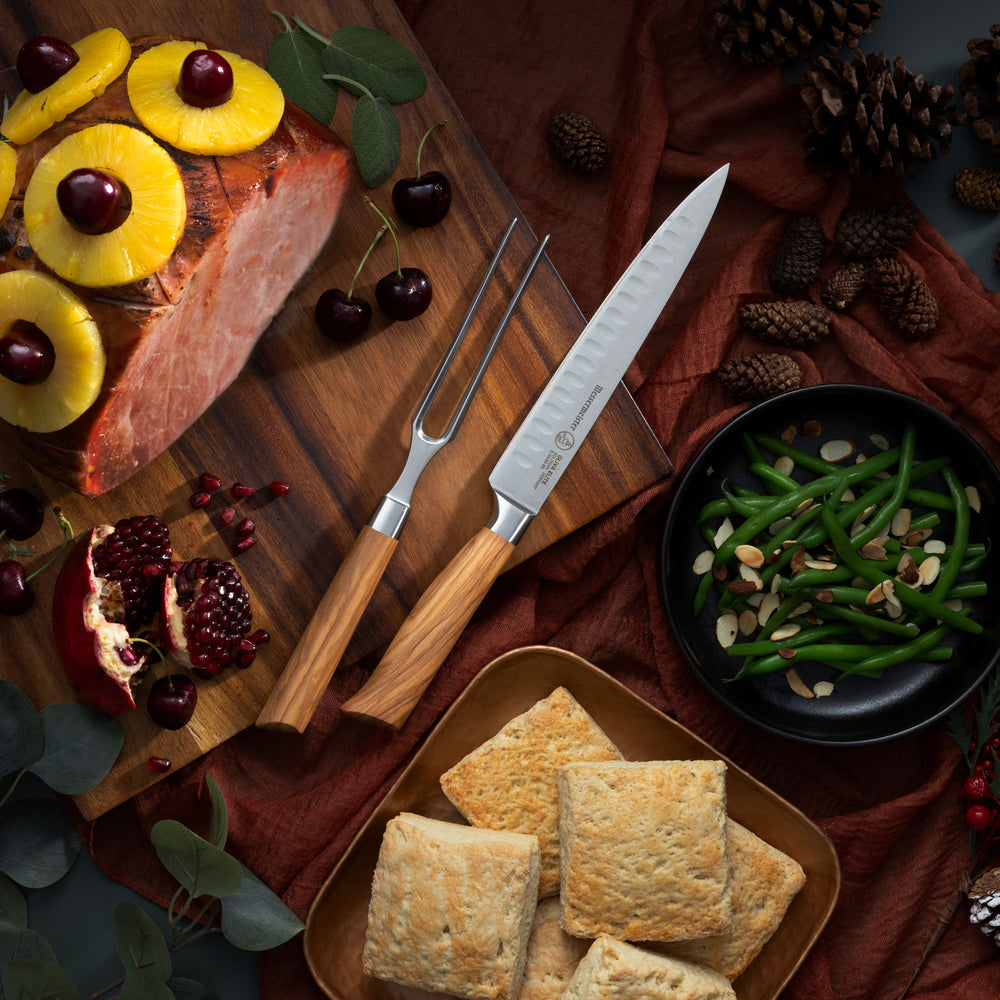 Wooden cutting board with sliced ham, pineapple, and cherries, accompanied by green beans and biscuits featuring the Messermeister Oliva Carving Set with Slicing Knife and Fork.