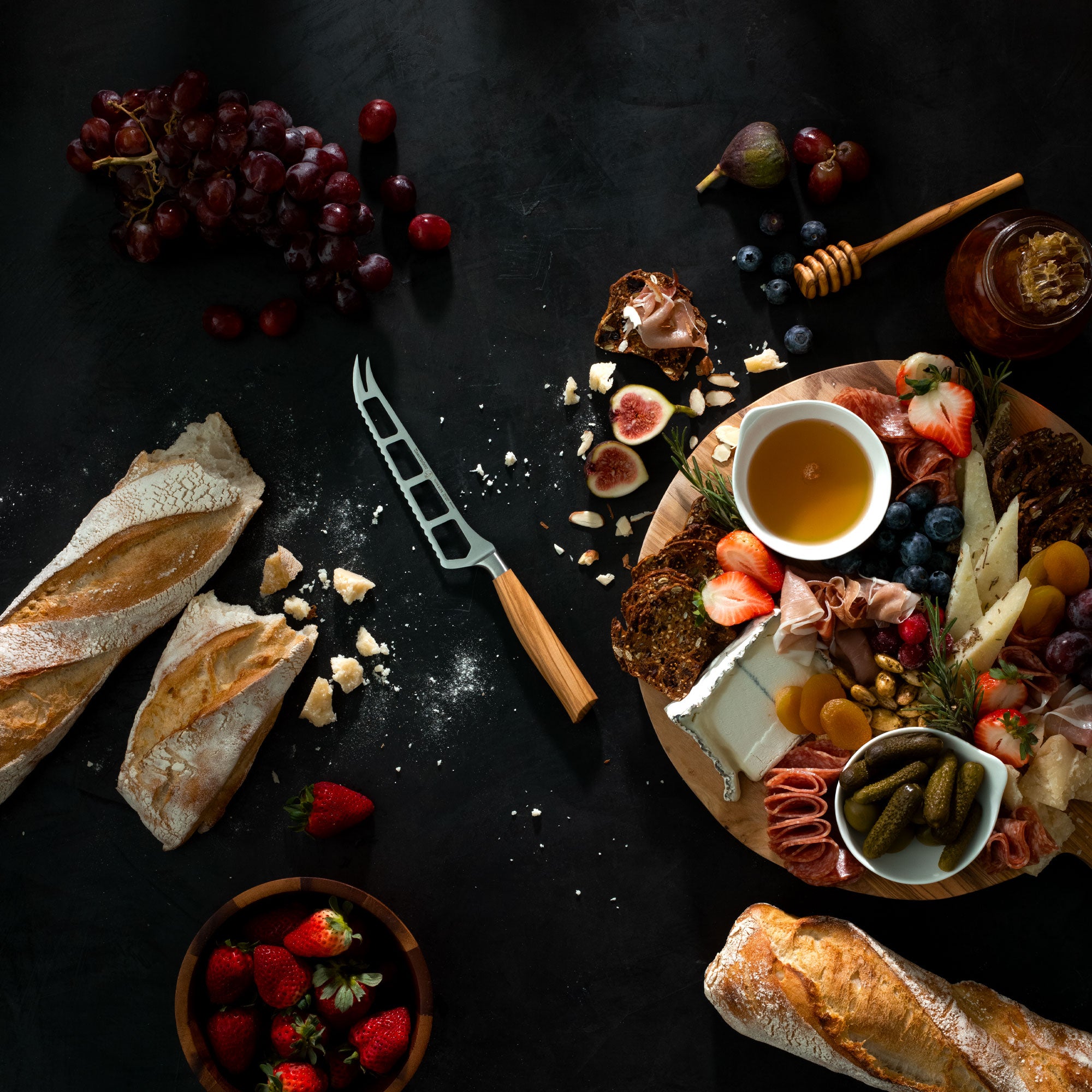 Charcuterie Platter of assorted meats, cheeses, bread, and fruits on a dark surface with a Messermeister Oliva Cheese and Tomato knife and honey dipper.
