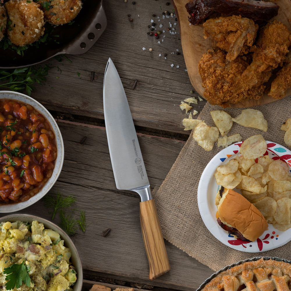 Assorted food items including a Messermeister Oliva Chef knife on a wooden table in a picnic setting