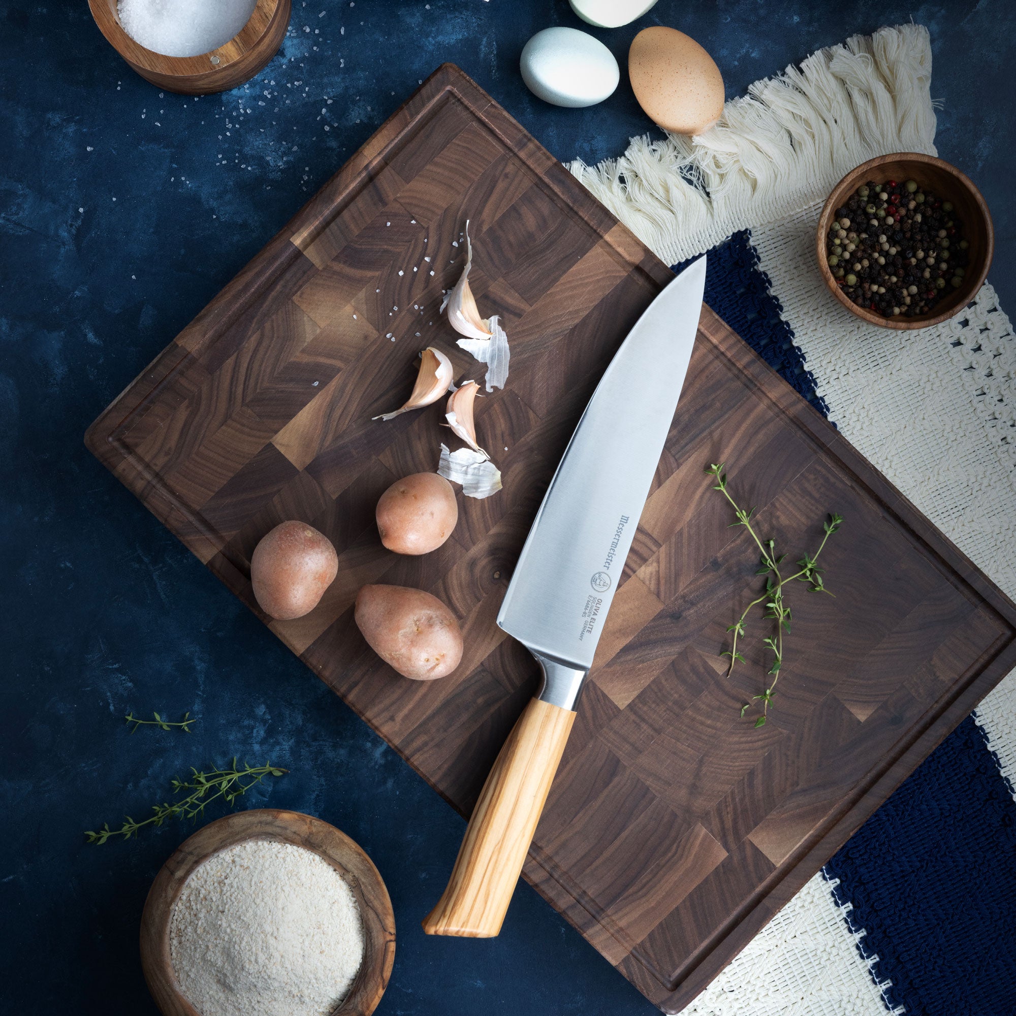 Wooden cutting board with Messermeister Oliva Chef knife, potatoes, garlic, and herbs on a dark surface