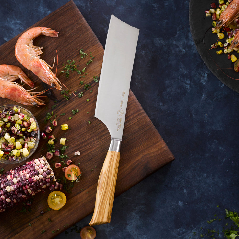Messermeister Oliva Nakiri Knife on a wooden cutting board with shrimp, corn, and vegetables on a dark surface