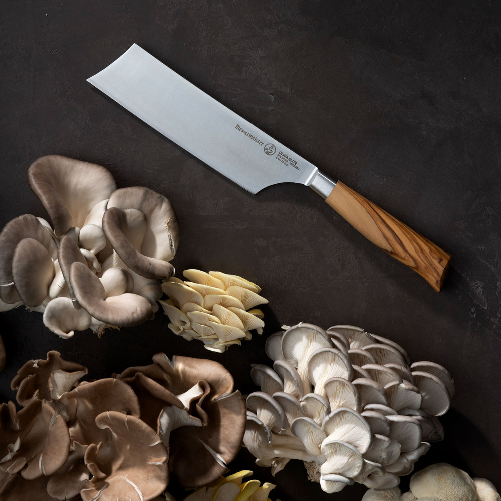 Messermeister Oliva Nakiri Knife with wooden handle on a dark surface surrounded by various mushrooms