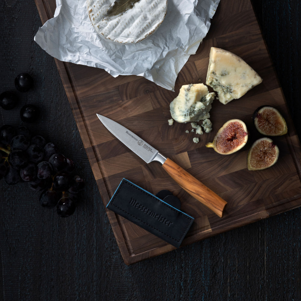 Wooden cutting board with cheese, figs, and a Messermeister Oliva Paring knife and Navy Magnetic Leather Edge Guard on a dark surface