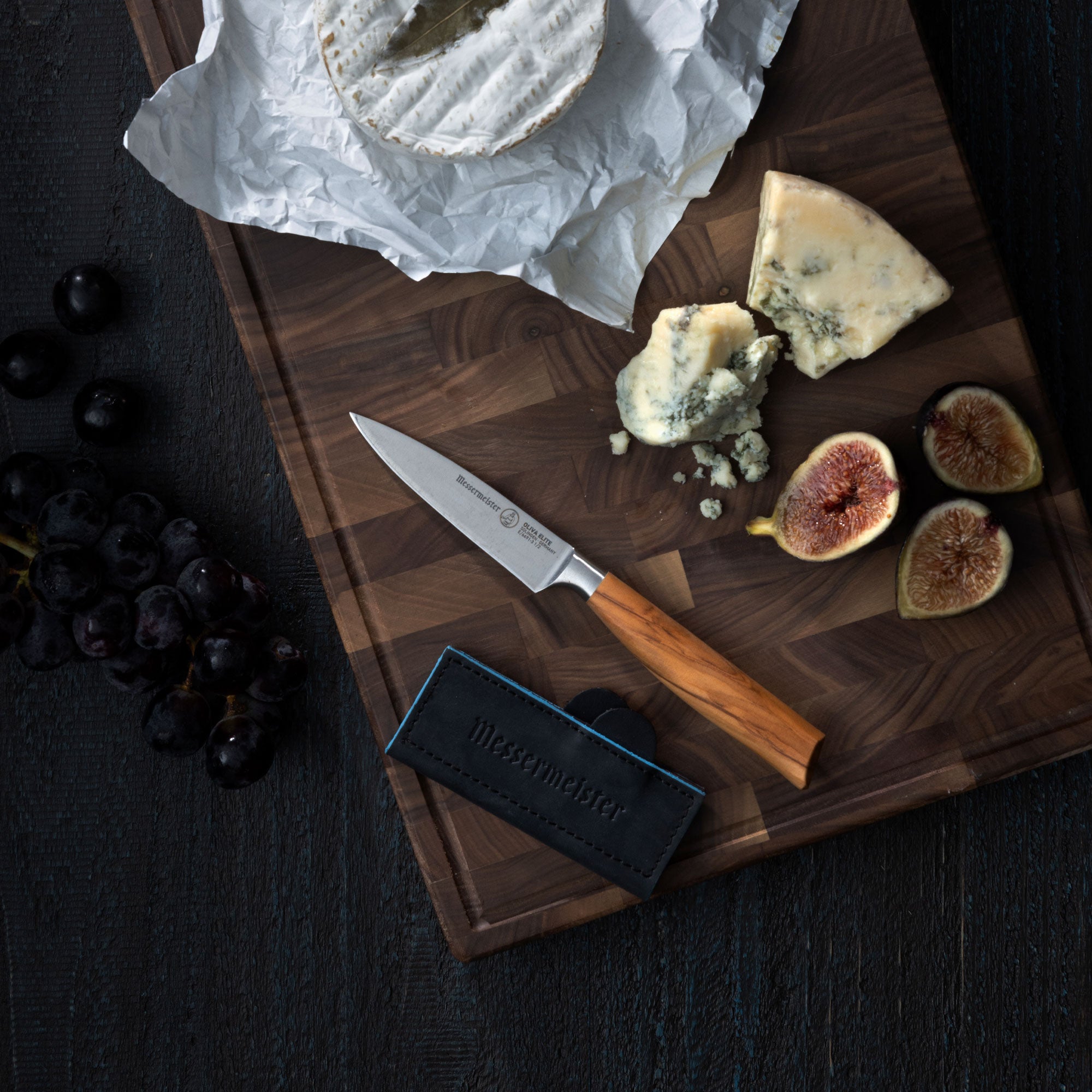 Wooden cutting board with cheese, figs, and a Messermeister Oliva Paring knife and Navy Magnetic Leather Edge Guard on a dark surface