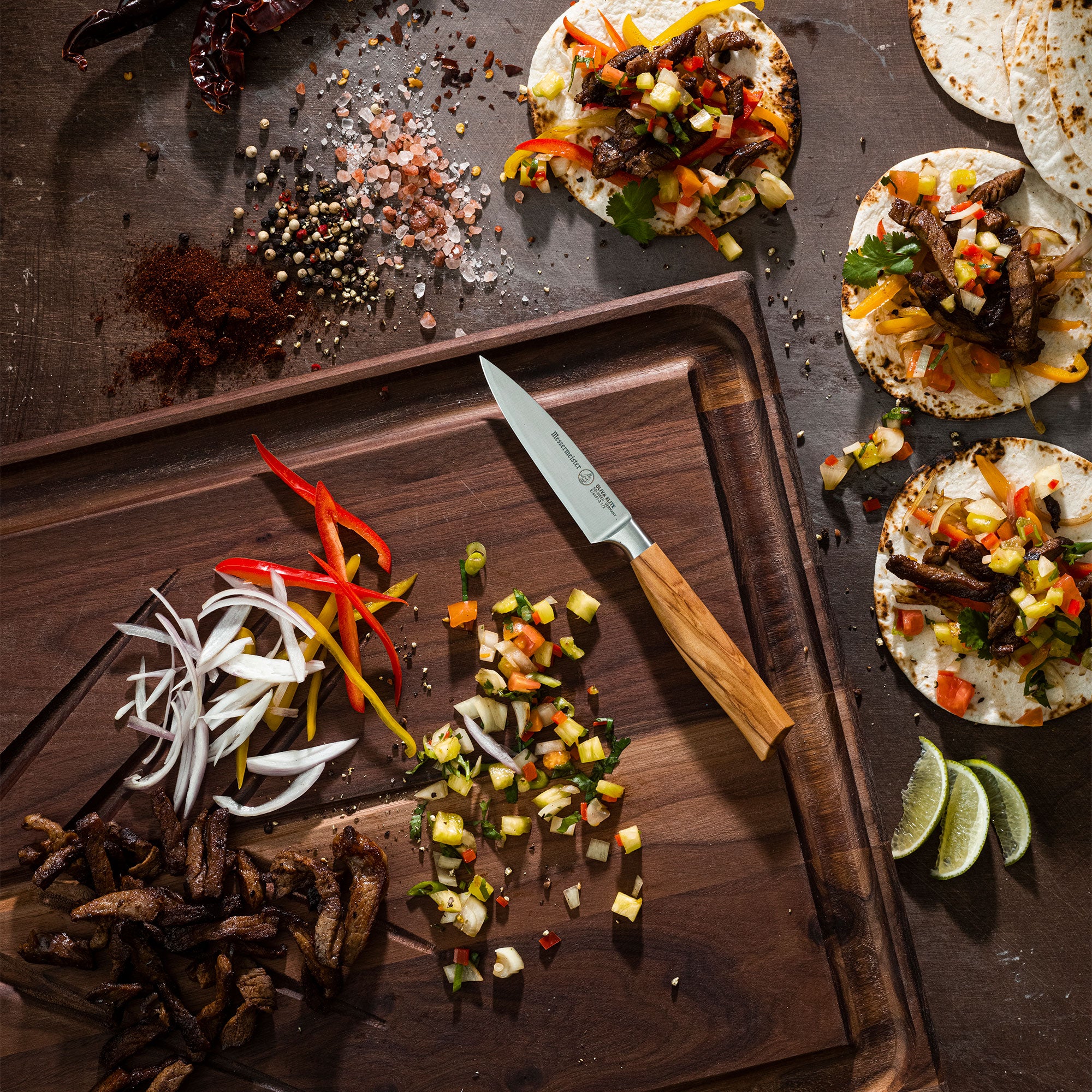 Wooden cutting board with sliced vegetables, a Messermeister Oliva Paring knife, and tortilla pizzas on a dark surface.