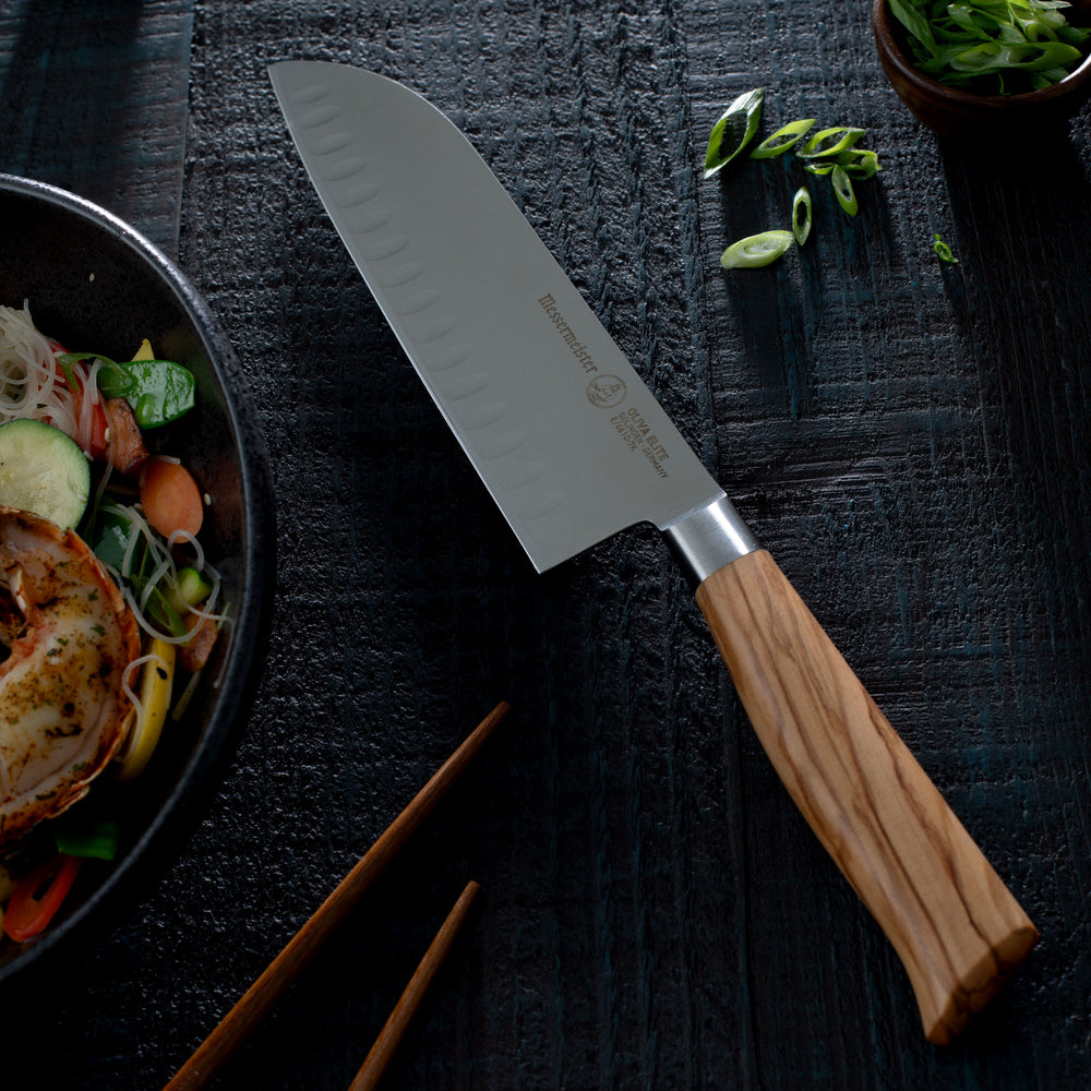 Messermeister Oliva Santoku knife with wooden handle on a dark surface next to a stir fry and chopsticks