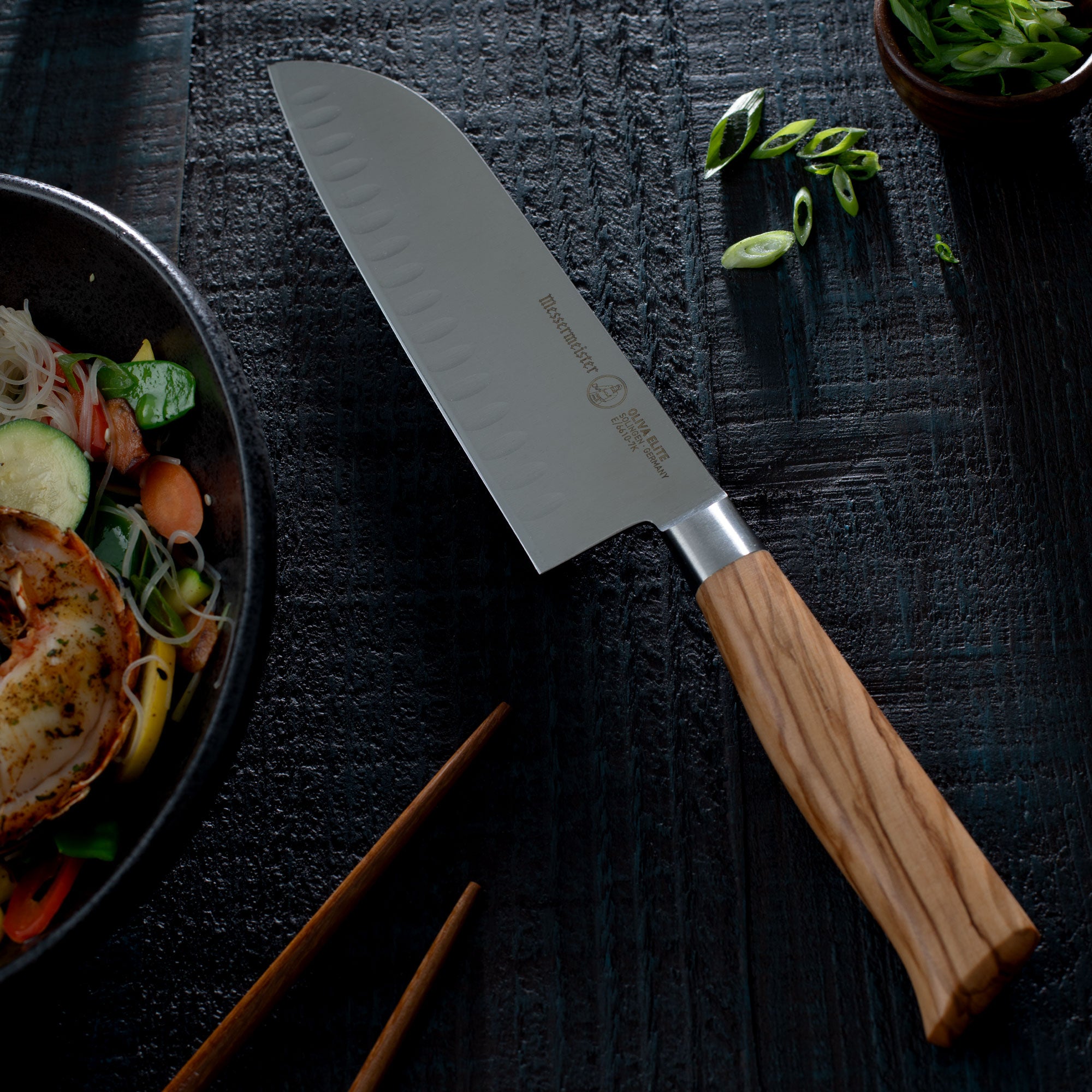 Messermeister Oliva Santoku knife with wooden handle on a dark surface next to a stir fry and chopsticks