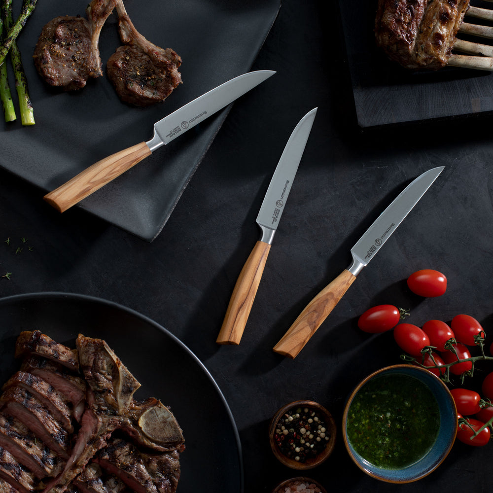 Messermeister Oliva Steak knives with Olive wood handles on a dark surface with grilled meat and vegetables.