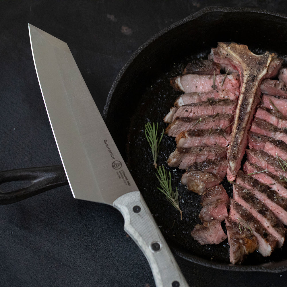 Sliced steak in a cast iron skillet with a Overland Chef Camp Knife on a dark surface