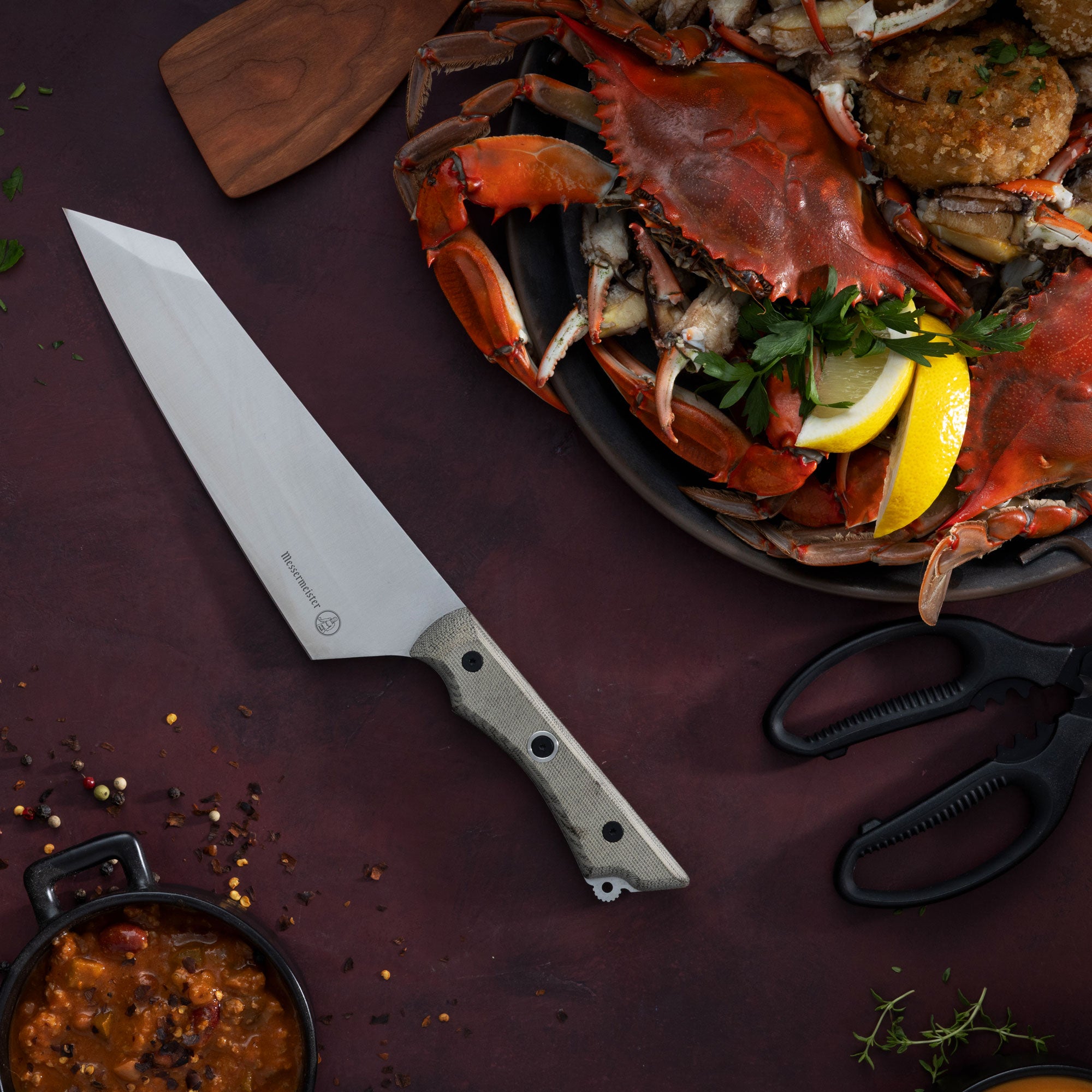 Messermeister Overland Chef Camp Knife on a dark surface with a platter of seafood including a crab and lemon wedges.