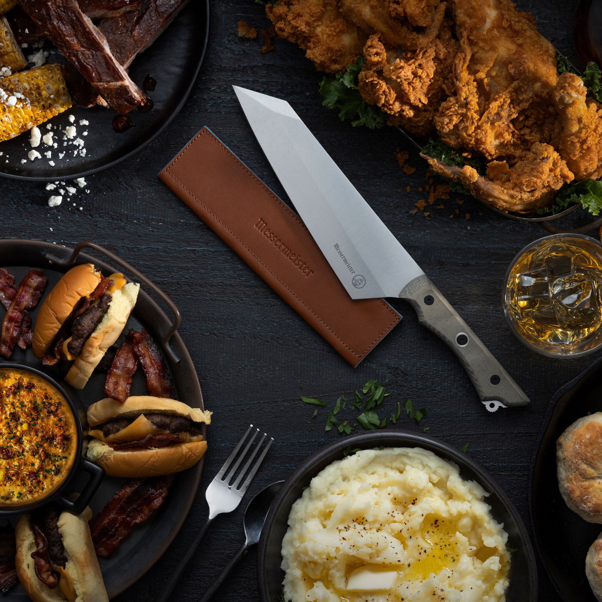 Assorted food items including burgers, fries, and mashed potatoes with Messermeister Overland Chef Camp 
knife on a dark surface with Messermeister Magnetic Leather Edge Guard Saddle Color.