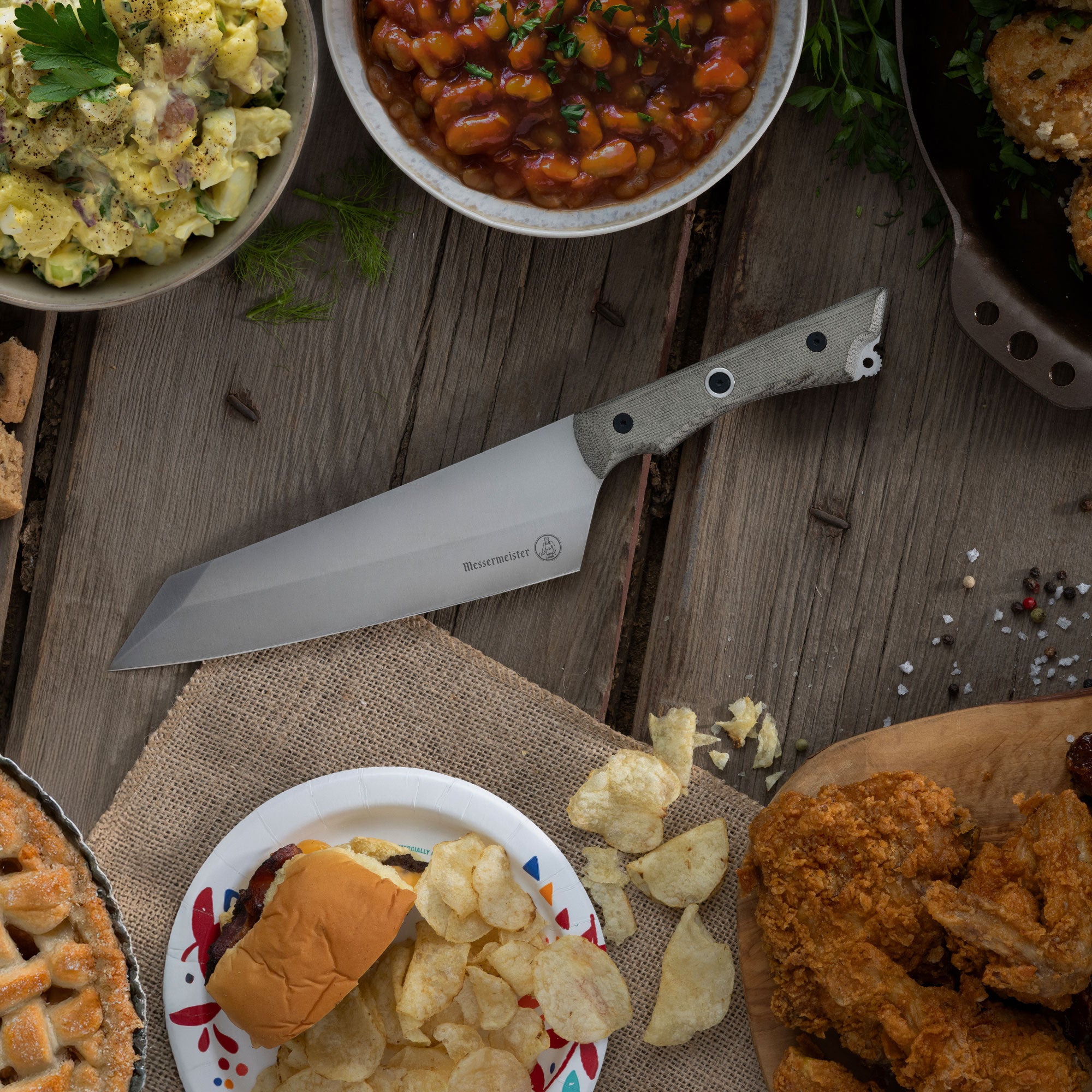 Messermeister Overland Chef Camp Knife on a wooden table with food including pie, chips, and beans.