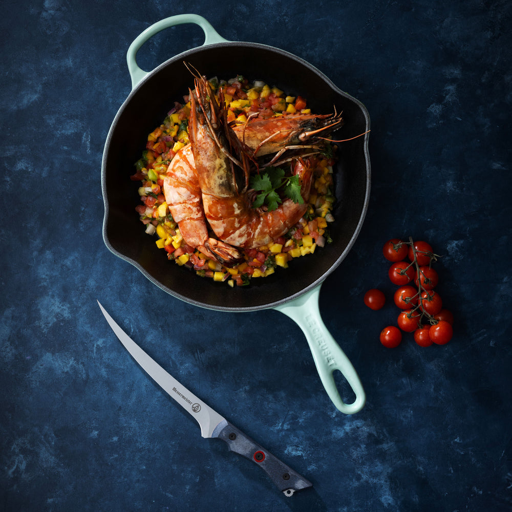 Messermeister Overland Chef Field and Flame Fillet Knife pictured with Prawns and Vegetable Medley in a Cast Iron Pan on a dark blue surface.