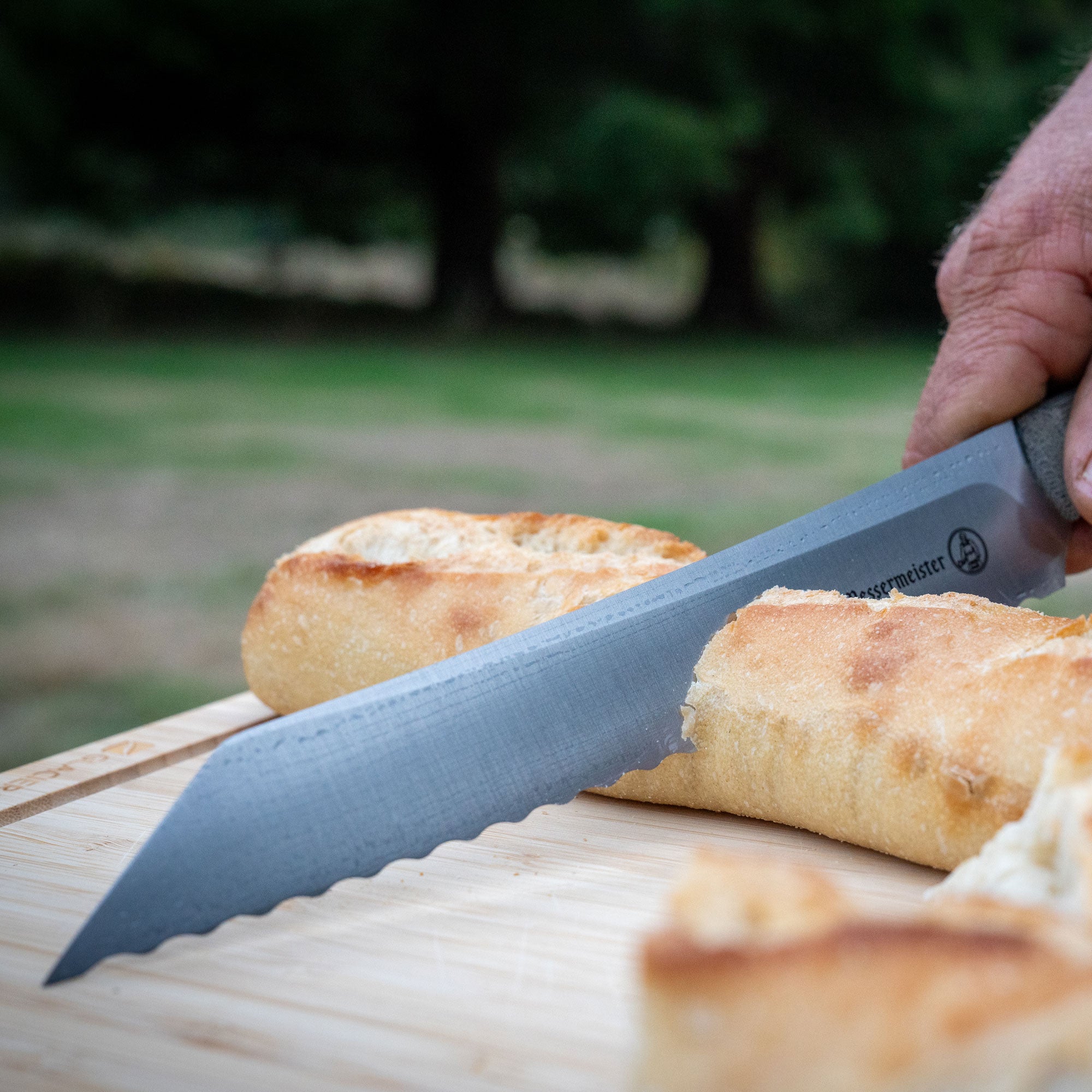 Person slicing bread with a Messermeister Overland Scalloped Slicing knife on a wooden board outdoors.