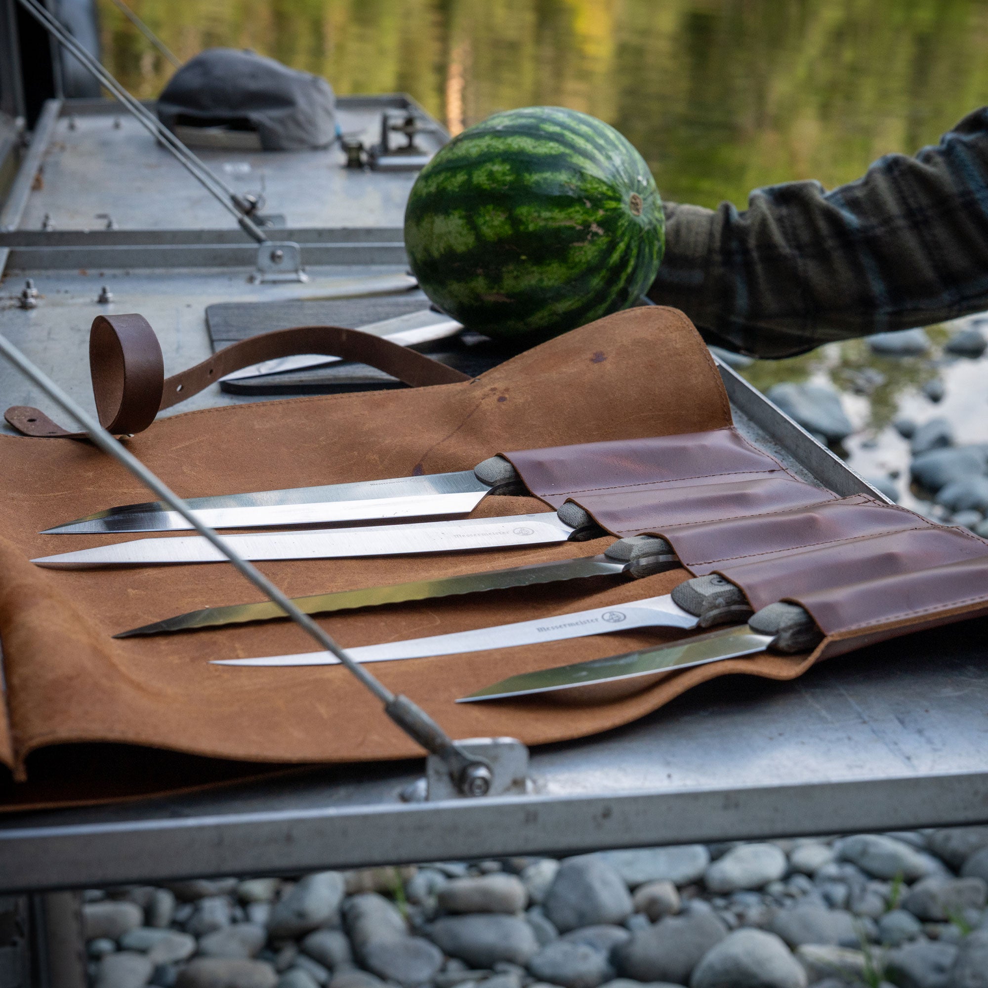 Messermeister Overland Chef Set of knives in a leather sheath with a watermelon on a metal surface near water.