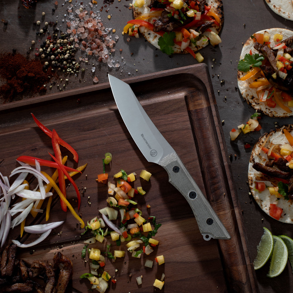 Messermeister Overland Chef Utility Knife on a wooden cutting board with chopped vegetables and tortilla pizzas.