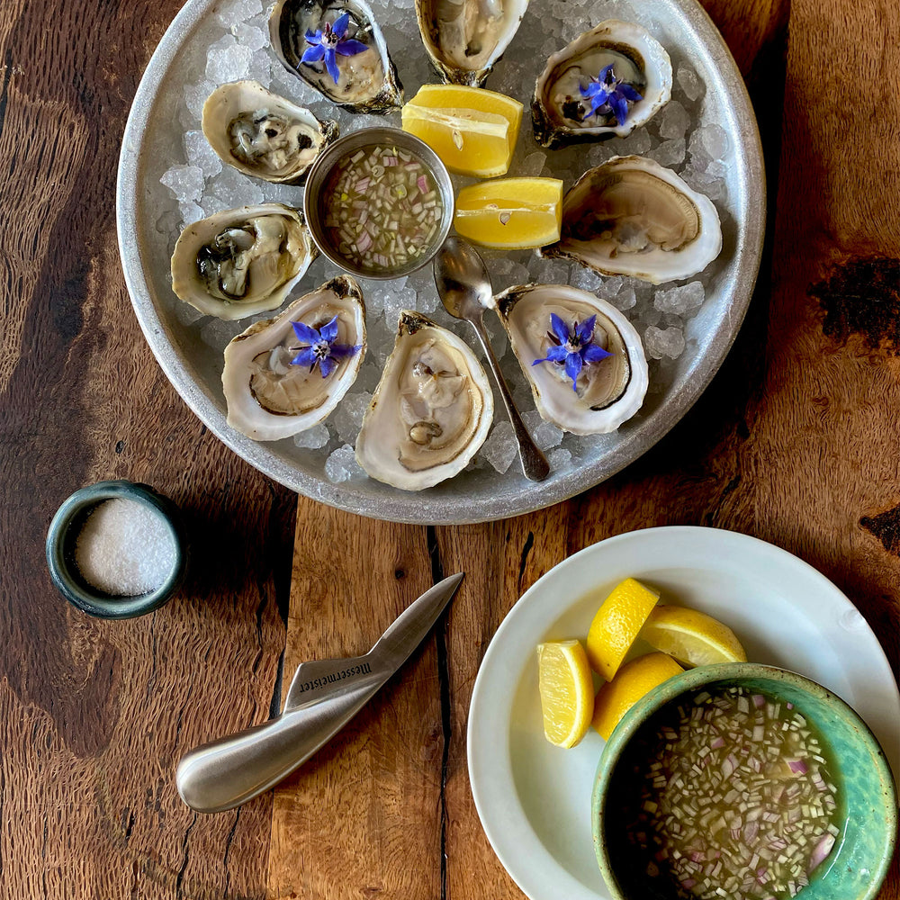Platter of oysters on ice with lemon wedges and a small bowl of sauce with Messermeister Oyster Shucking Knife on a wooden surface.