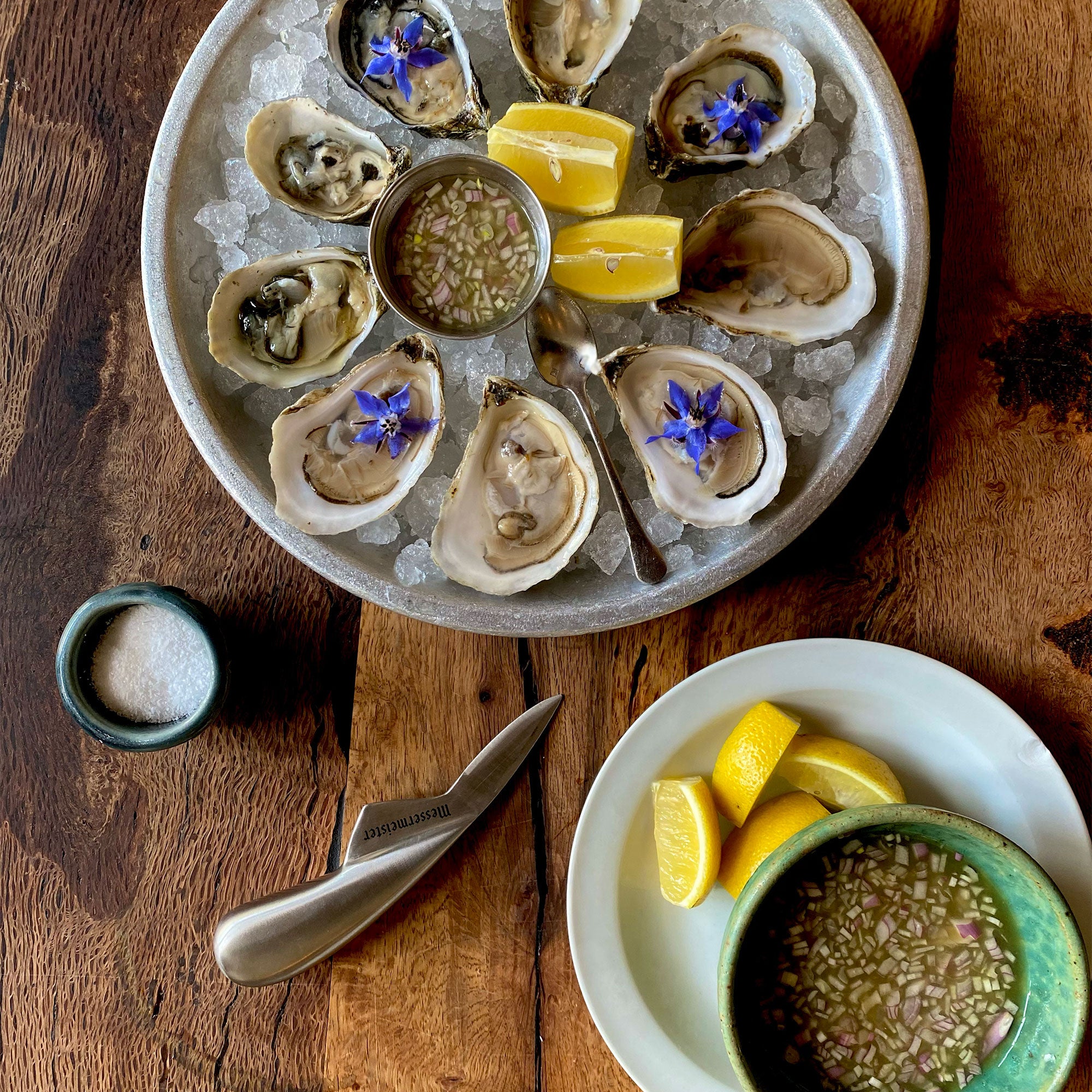 Platter of oysters on ice with lemon wedges and a small bowl of sauce with Messermeister Oyster Shucking Knife on a wooden surface.