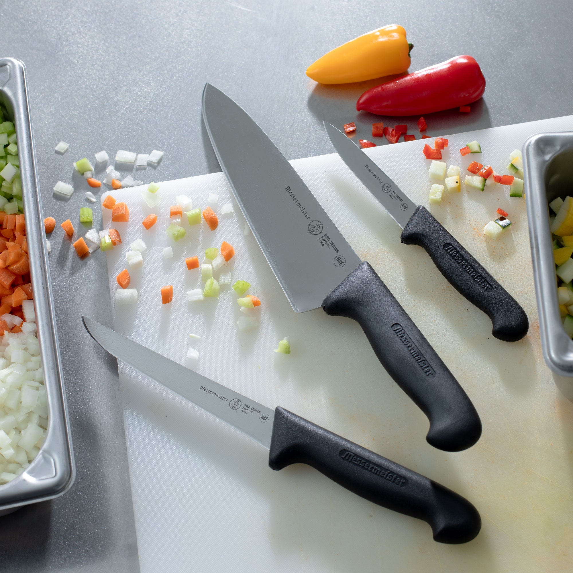 Messermeister Pro Series Chef, Utility and Paring Knife on a cutting board with chopped vegetables on a stainless steel prep kitchen table.