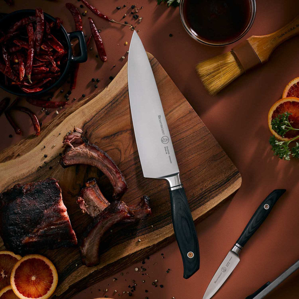 Messermeister Blacksmith 8-Inch Chef’s Knife with a hammered high-carbon stainless steel blade and ebony pakkawood handle resting on a rustic wood cutting board beside smoky BBQ ribs, styled for outdoor grilling and premium kitchen craftsmanship