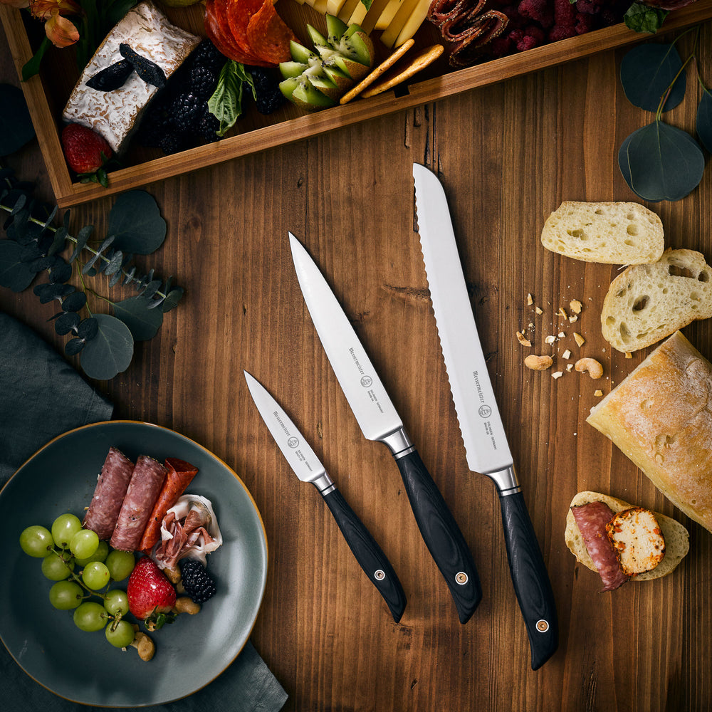 Messermeister Blacksmith knife set including bread, utility, and paring knives with hammered high-carbon stainless steel blades and ebony pakkawood handles, arranged on a rustic wood cutting board beside fresh bread and a charcuterie spread for artisanal kitchen presentation