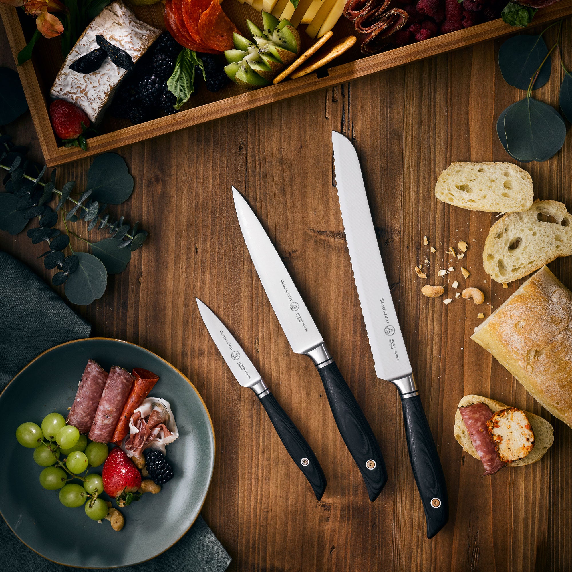 Messermeister Blacksmith knife set including bread, utility, and paring knives with hammered high-carbon stainless steel blades and ebony pakkawood handles, arranged on a rustic wood cutting board beside fresh bread and a charcuterie spread for artisanal kitchen presentation