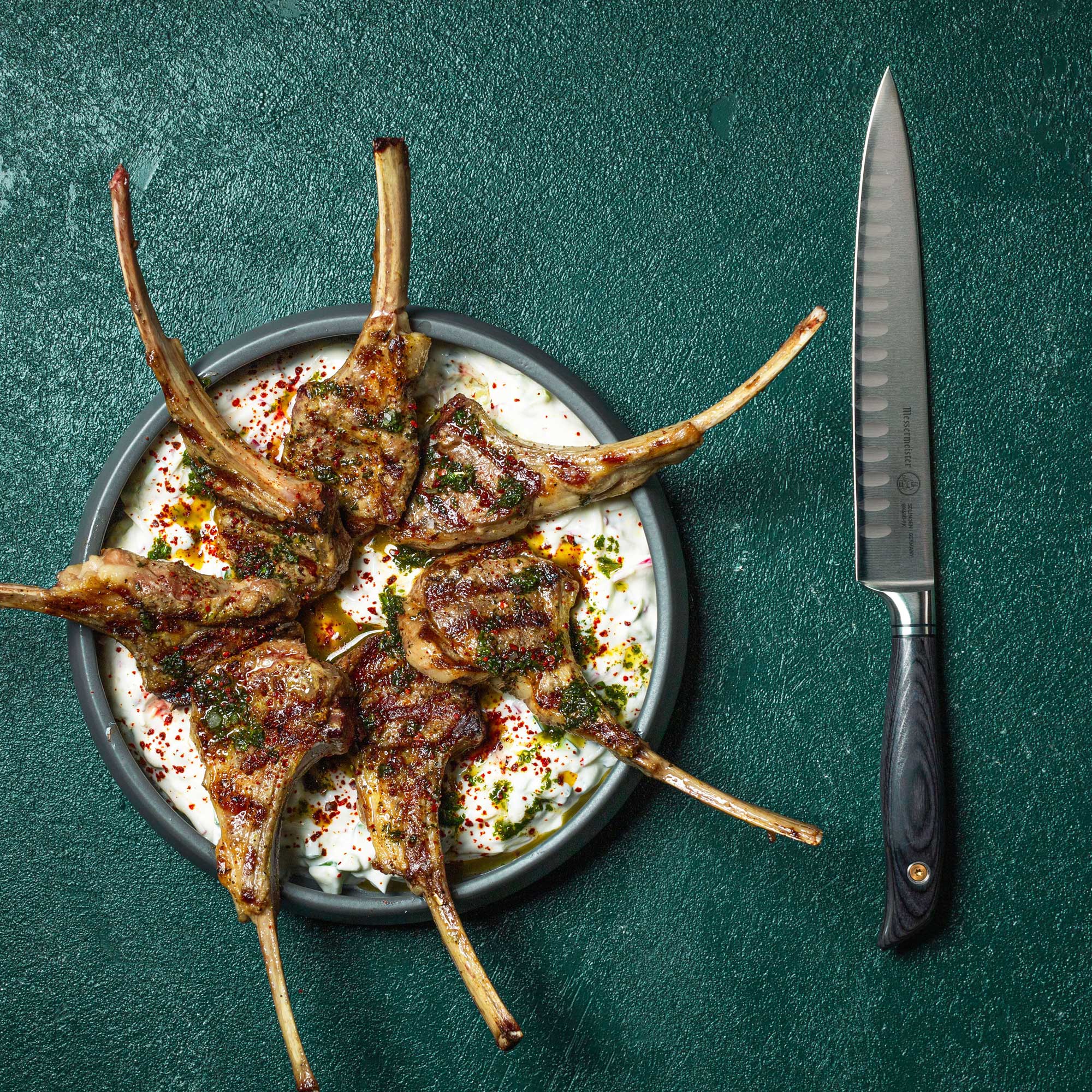 Messermeister Blacksmith Kullens  Carving Knife - 9" with stainless steel blade and ebony pakkawood handle- on a green table next to a plate of delicious looking lamb chops