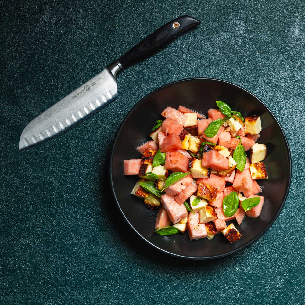 Messermeister Blacksmith Kullens Santoku Knife - 6.5" - on a green table next to a plate of diced chicken and watermelon