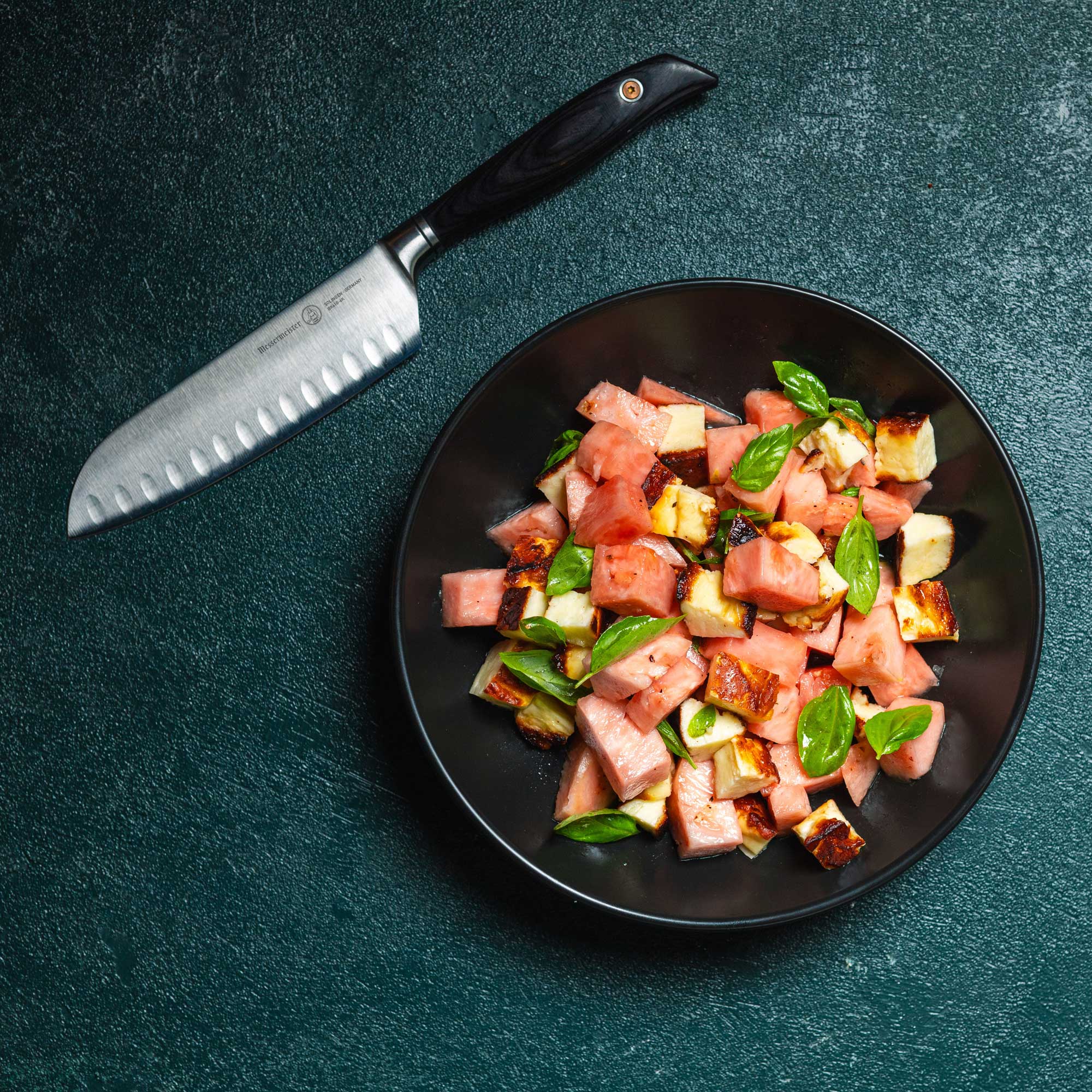 Messermeister Blacksmith Kullens Santoku Knife - 6.5" - on a green table next to a plate of diced chicken and watermelon