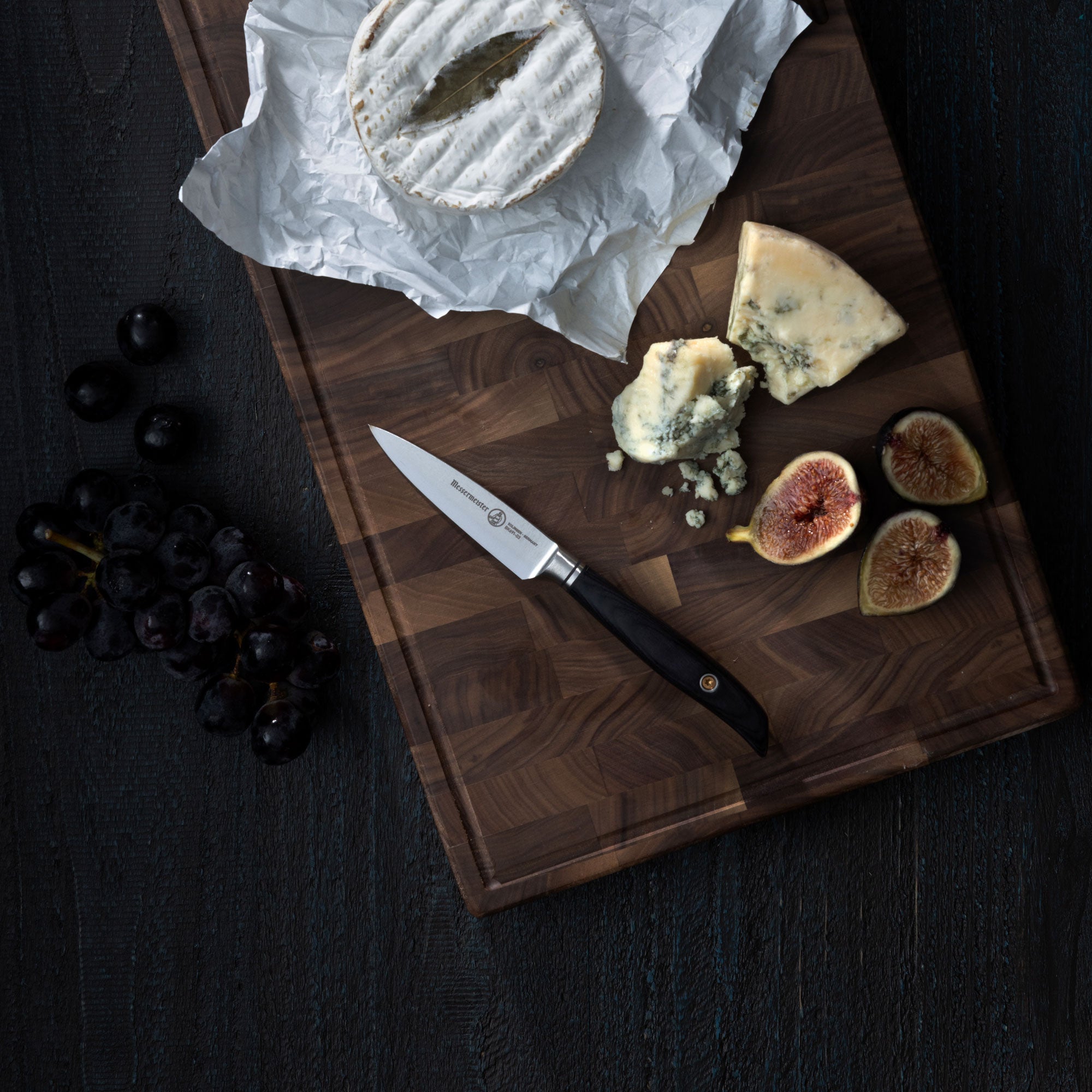 Messermeister Blacksmith Paring Knife on a cutting board with cheese and figs that lay on a dark wood surface with grapes on it. 