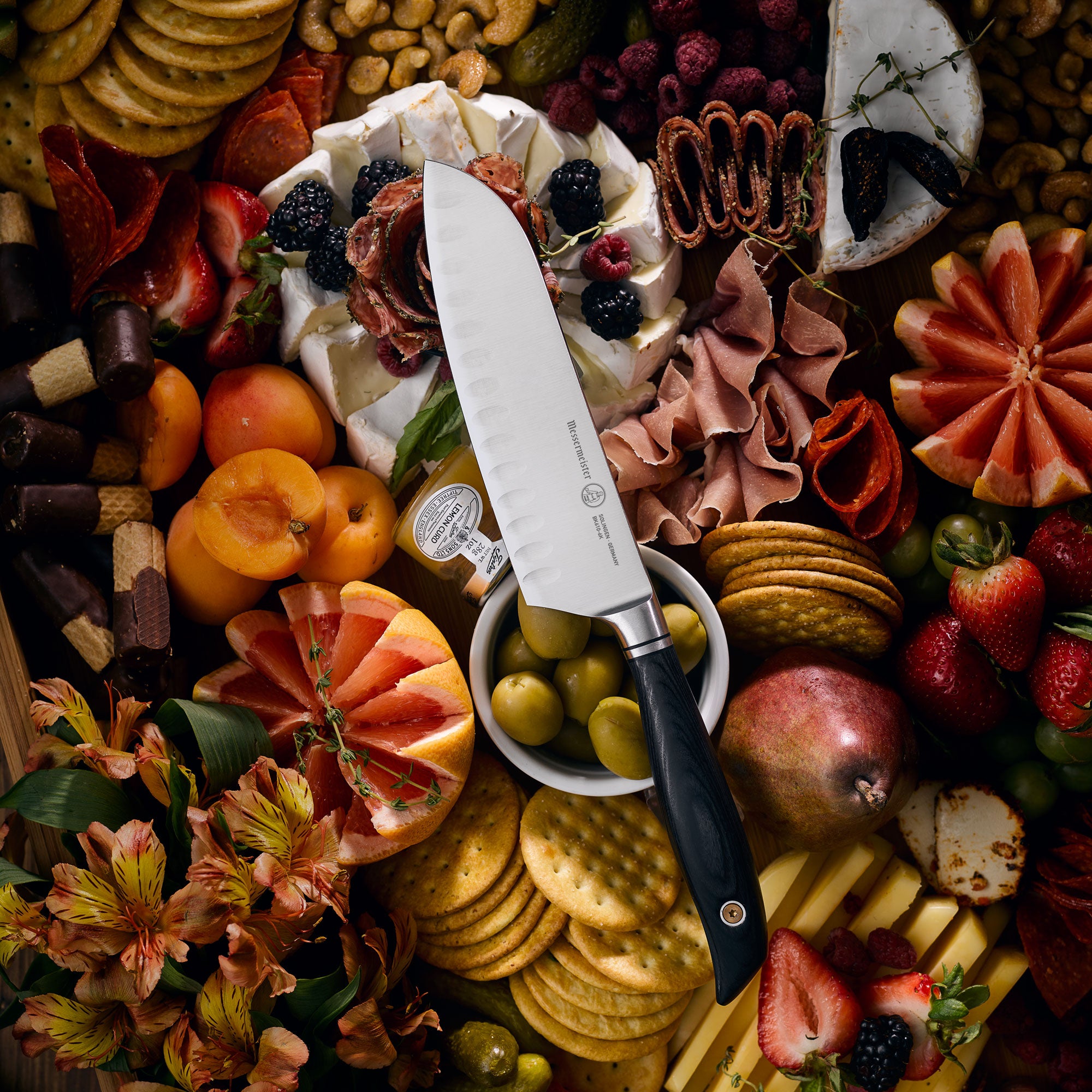 Messermeister Blacksmith Santoku Knife resting on a charcuterie board surrounded by sliced cured meats, artisan cheeses,  crackers, fresh herbs, and seasonal fruits, highlighting the knife’s satin-finish blade and blue handle