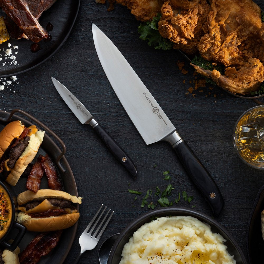 Assorted food items including burgers, fries, and mashed potatoes on a dark surface with a Messermeister Blacksmith Chef's Knife and Paring Knife and a glass of bourbon.