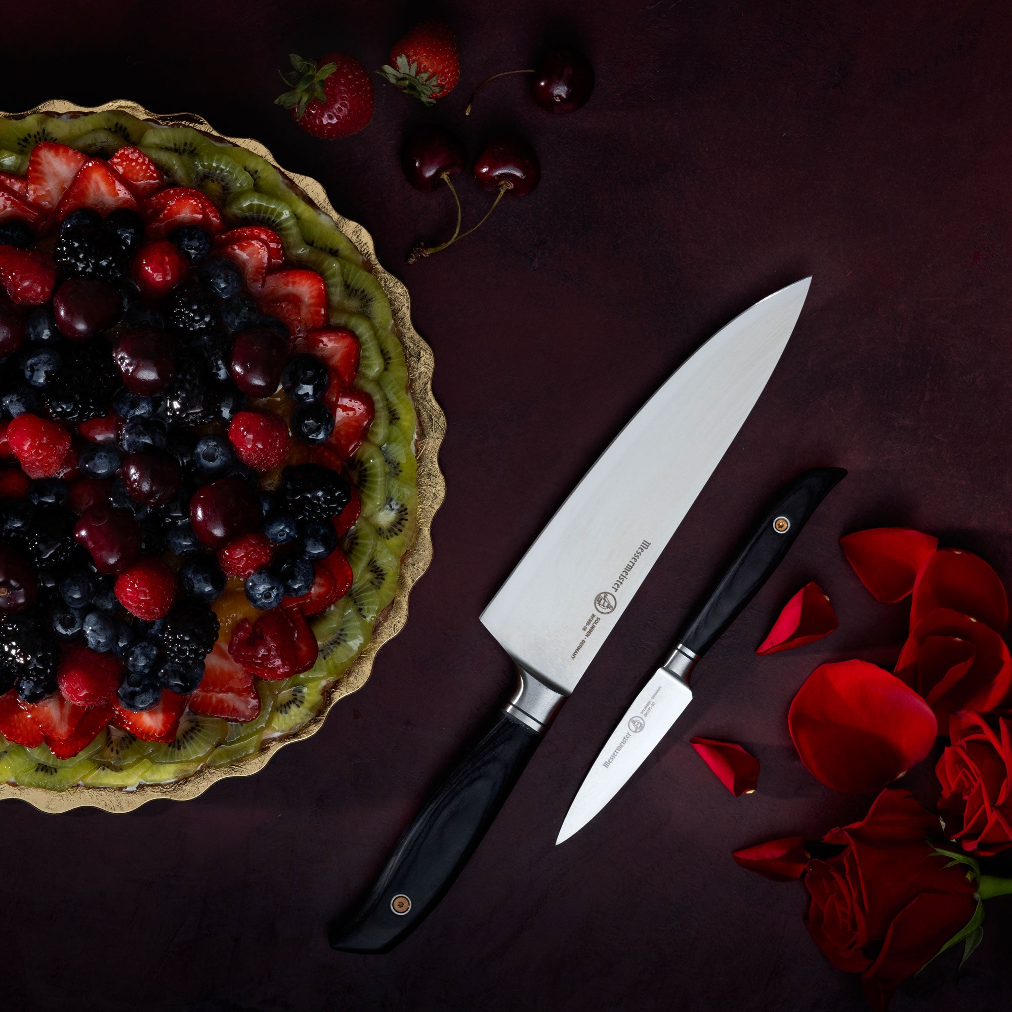 Messermeister Blacksmith Chef Knife and Parer with a fruit tart, cherries, strawberries and rose petals on a dark red surface