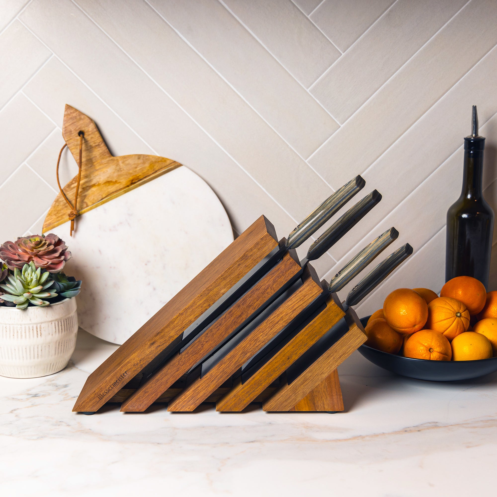Messermeister Carbon 5 Piece Next Level Block Set shown with knives resting inside of knife block on a counter with oranges and succulents in the background