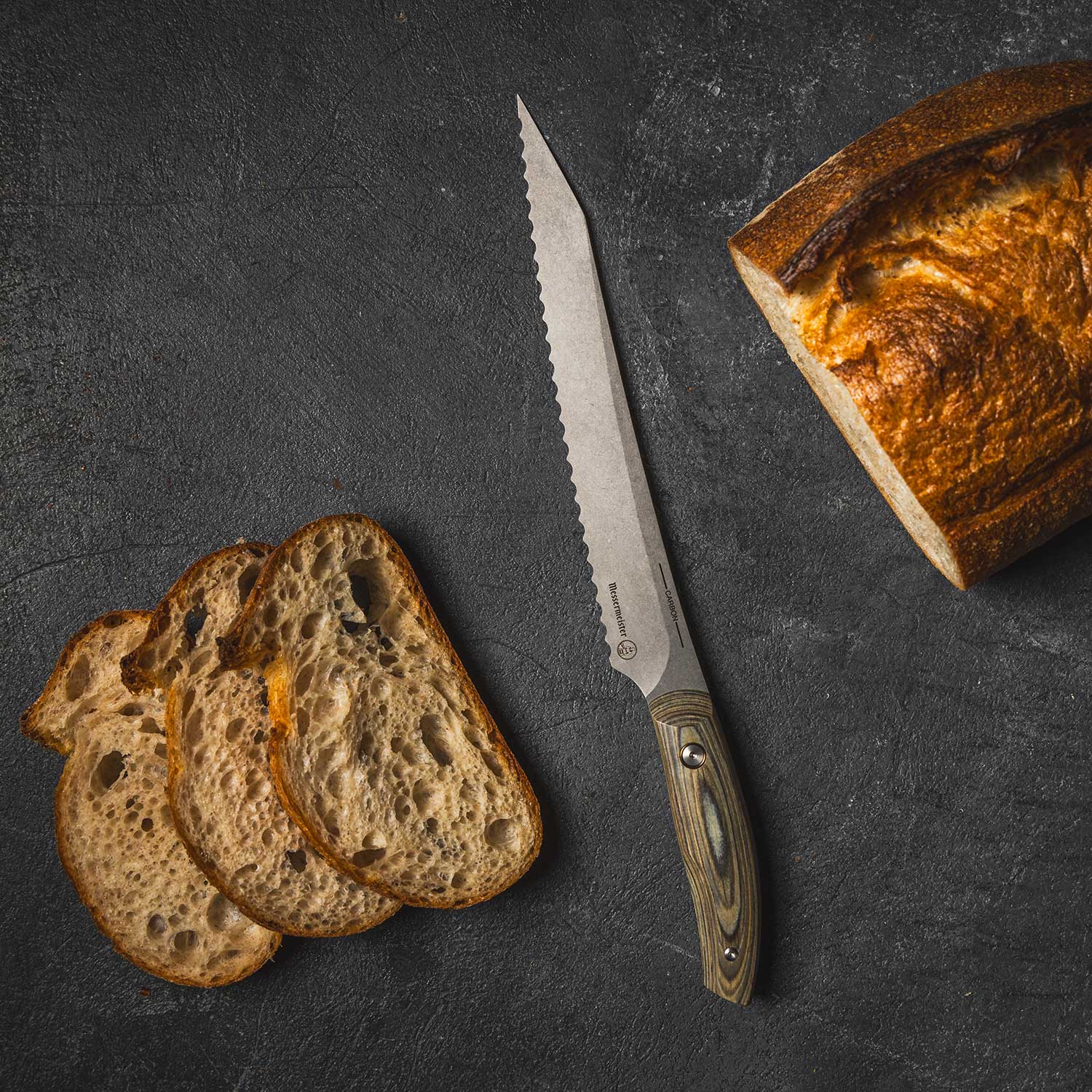 Messermeister 9 inch Carbon Bread Knife with serration and a Spanish hardwood handle lying next to a loaf of bread and pieces of sliced bread