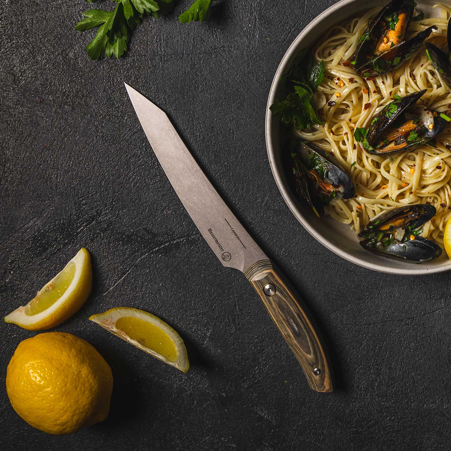 Messermeister 6 inch Carbon Utility Knife with a Spanish hardwood handle  shown lying beside a bowl of seafood and pasta with wedges of lemon