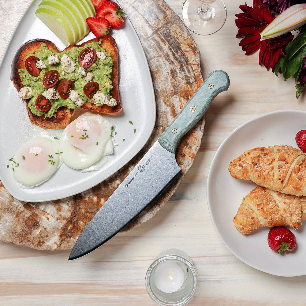 Messermeister Custom Blue 8-inch chef’s knife displayed beside pastries, avocado toast with eggs, and fresh fruit, featuring a  stainless steel blade and a bold blue ergonomic modular handle highlighting the sharp cutting edge