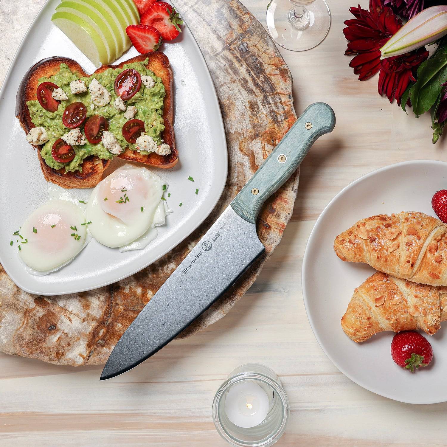 Messermeister Custom Blue 8-inch chef’s knife displayed beside pastries, avocado toast with eggs, and fresh fruit, featuring a  stainless steel blade and a bold blue ergonomic modular handle highlighting the sharp cutting edge